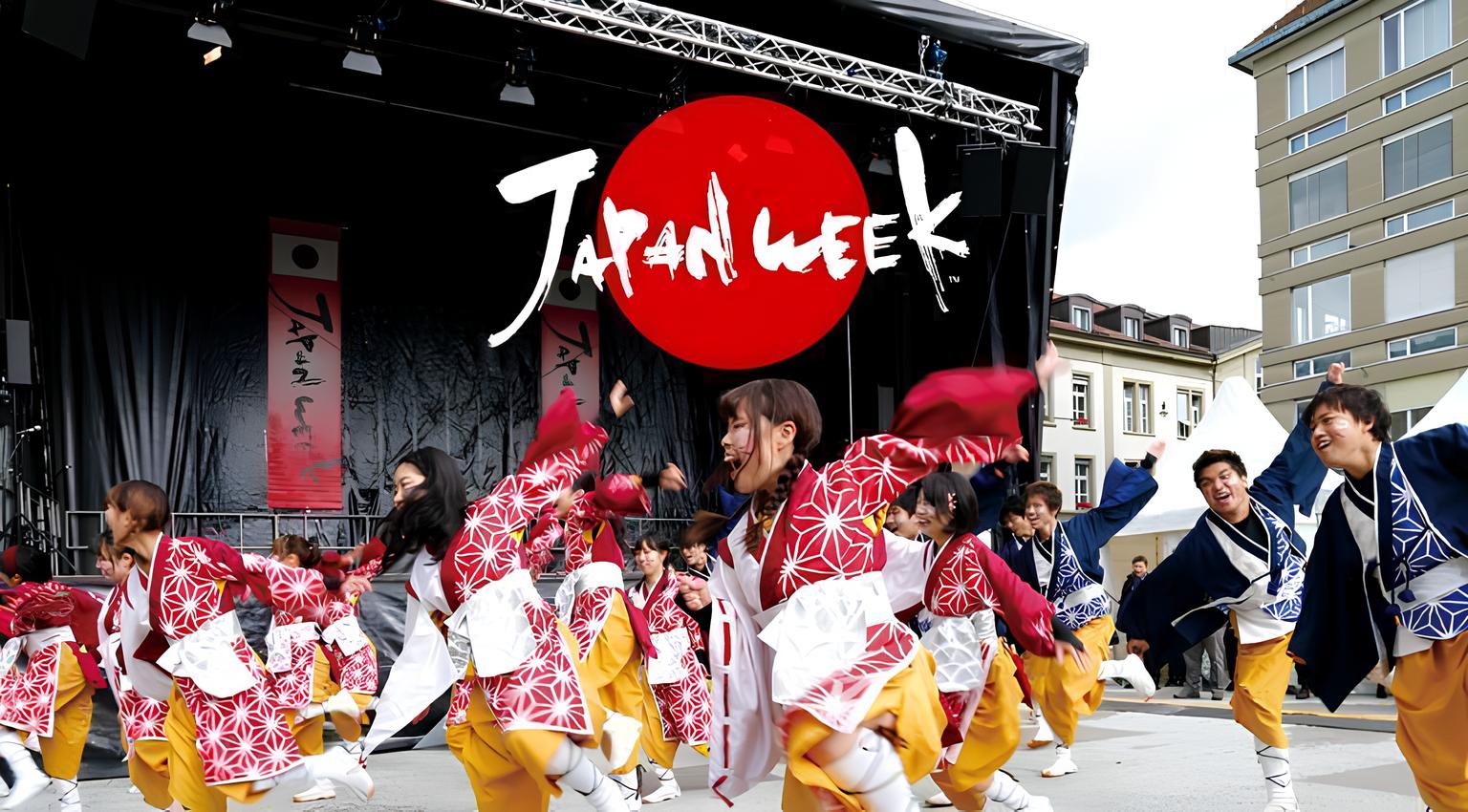 Group of dancers dressed in traditional Japanese clothes dancing outside in front of a stage that has the words 'Japan Week' written on a sign above it