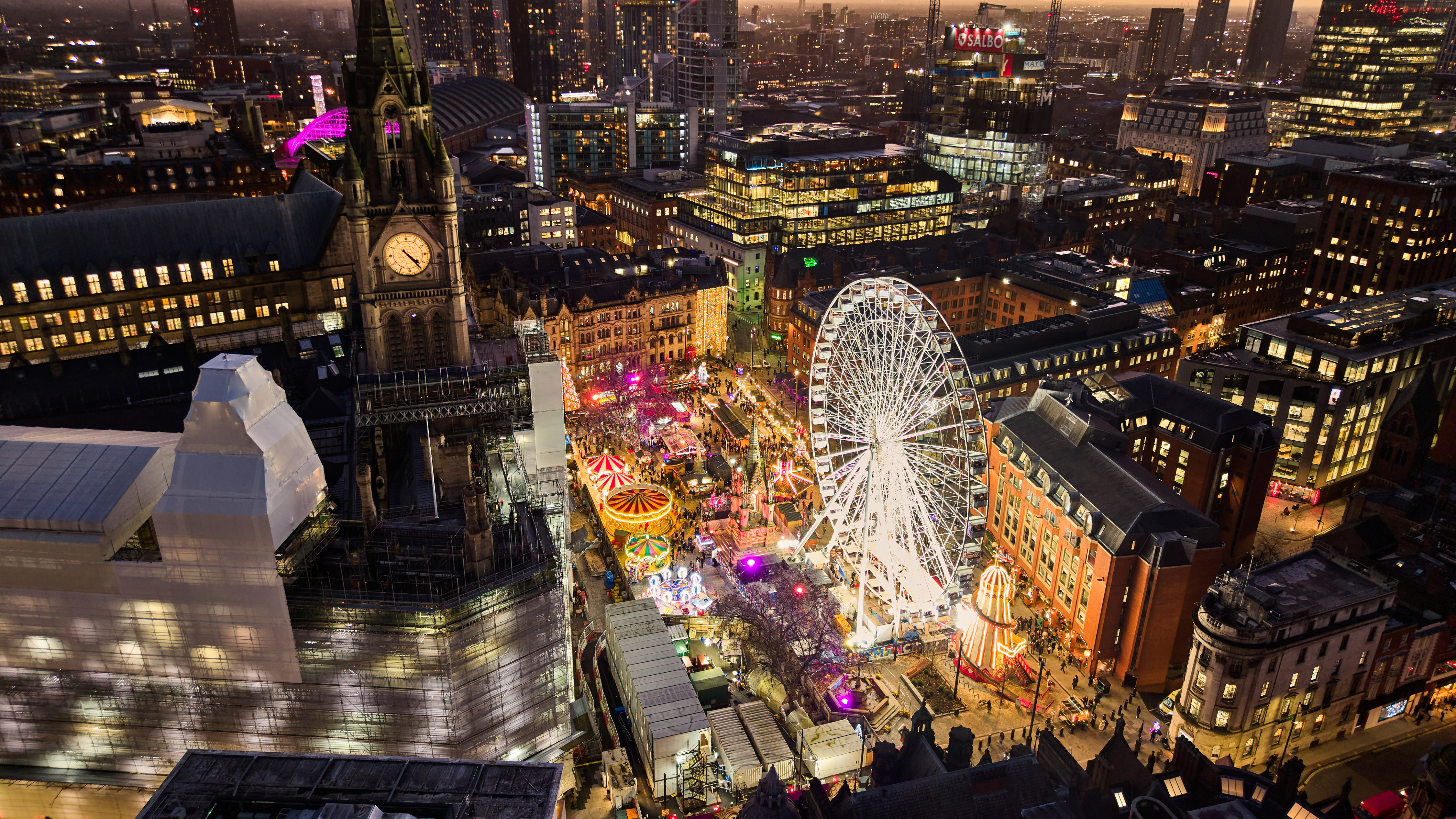Night time view across a city Manchester showing rooftops and buildings across the city including the clocktower of the Victorian town hall and a giant Ferris wheel lit up in white with fairground rides visible around it