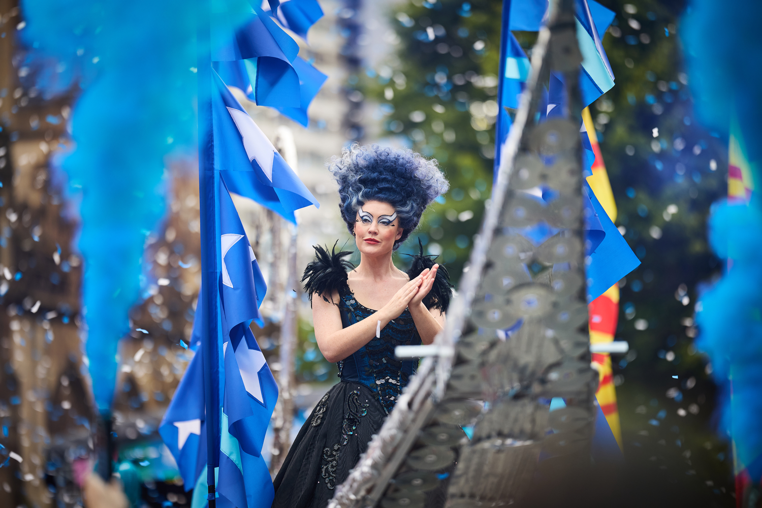 A woman wearing a theatrical high wig and black evening dress, standing atop a big silver gondola, with blue smoke cascading all around and coloured confetti raining down.