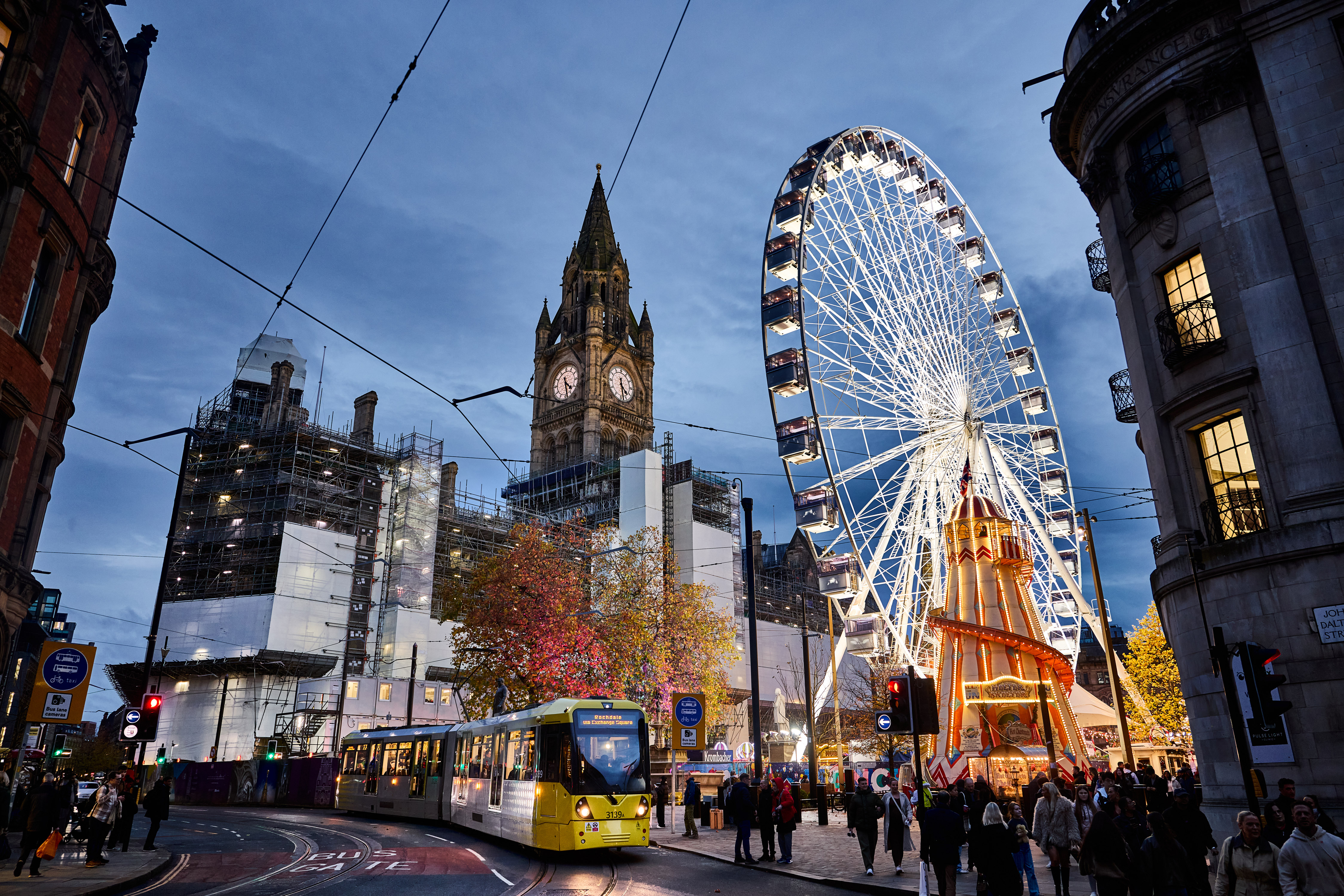 Street scene a bright yellow tram comes towards the camera passing by a square with a big building at the back of it and an enormous bright white Ferris wheel with a yellow and red Helter Skelter in front of it