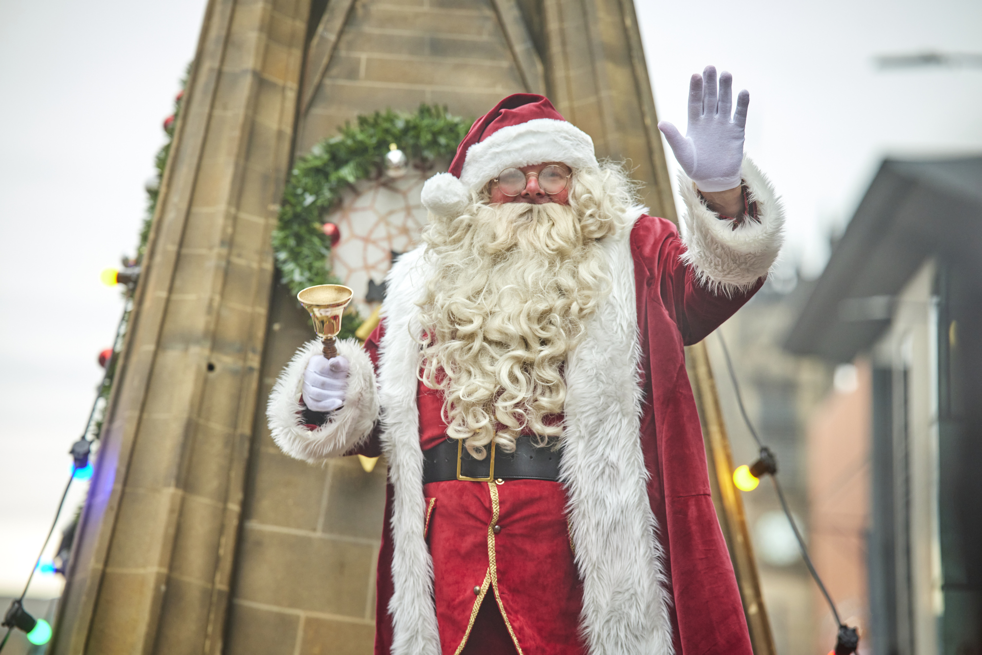 Father Christmas with a long white curly beard and moustache, wearing a red and white Santa suit and hat, ringing a handbell and waving