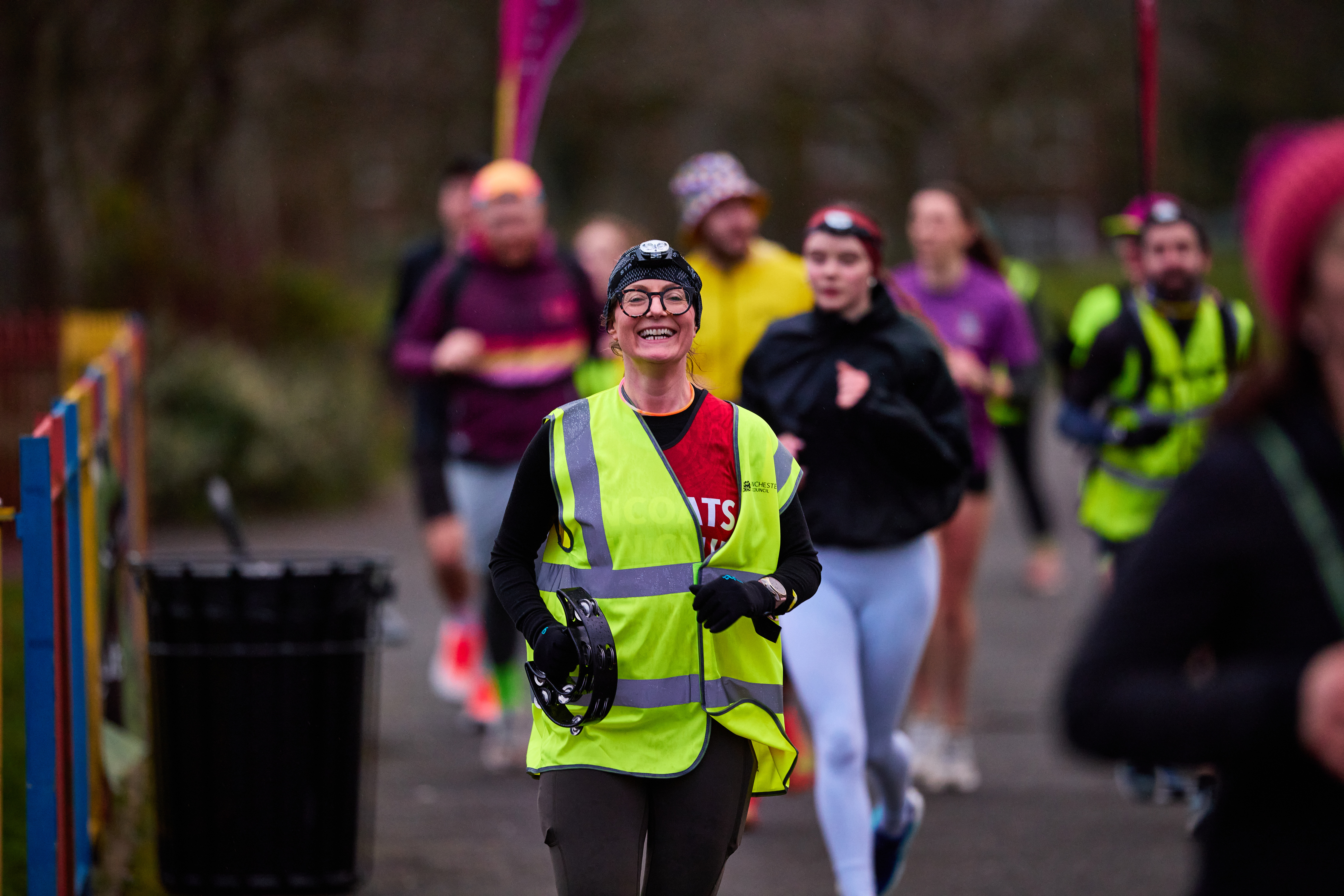 Outdoors a group of runners led by one runner wearing a yellow high-vis jacket and a woolley hat smiling as they run