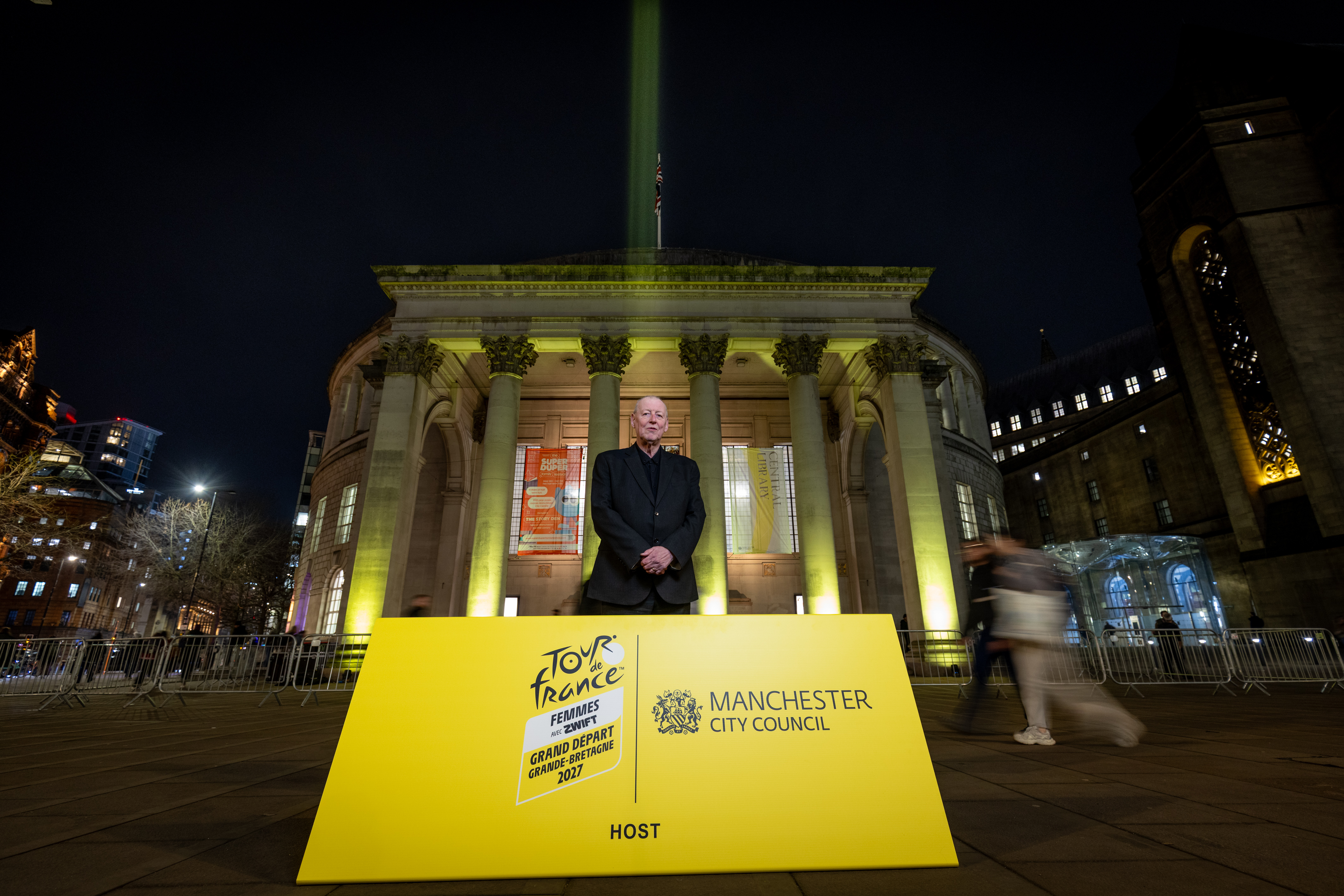 Man dressed in black standing in front of Manchester's Central Library which is lit up in yellow.  A big yellow sign can be seen in front of the man which reads Tour de France Femmes averc Zwift Grand Depart Grande Bretagne 2027 Manchester City Council