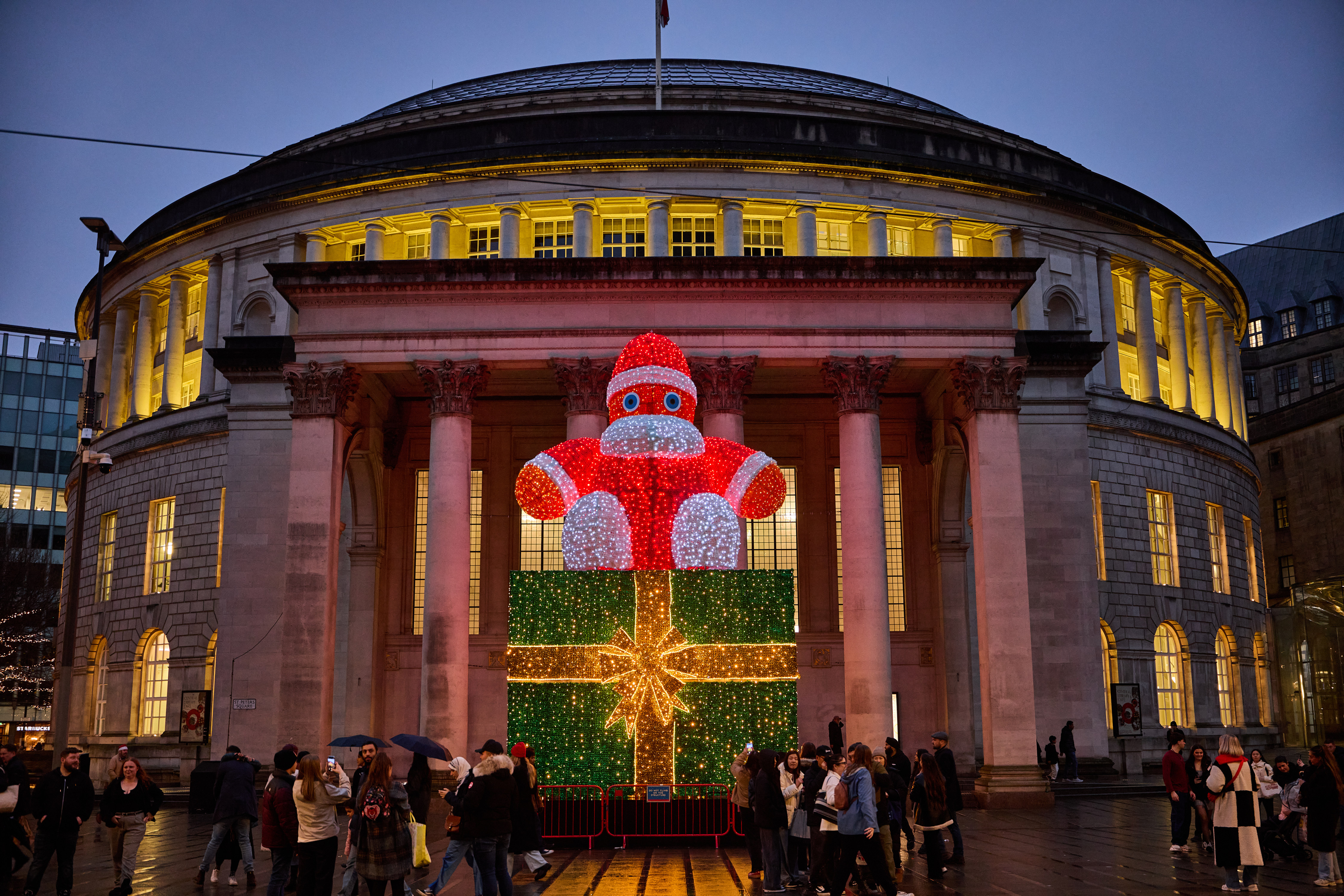 Street scene at night looking towards Manchester's iconic circular Central Library building with a big Santa figure in front of it that is sitting on top of a huge green square-shaped present - both are illuminated with twinkling lights