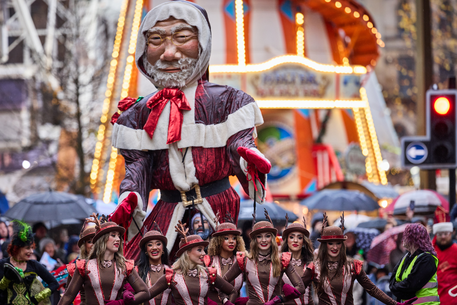 Parade street scene. Larger than life old Father Christmas puppet in brown with red ribbon round neck and white collar standing high above a troupe of dancers dressed as reindeer