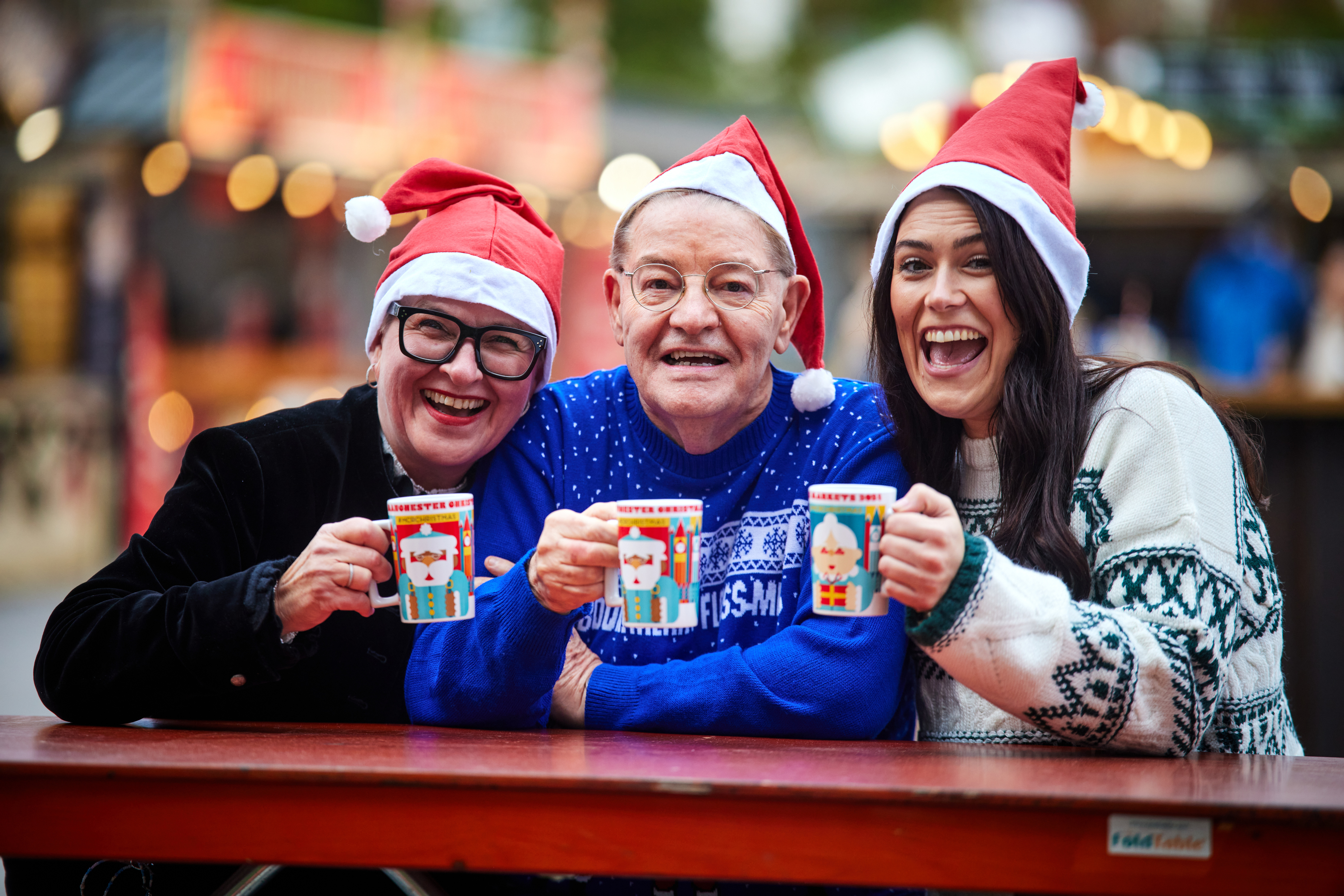 Two women and one man all wearing festive jumpers and Santa hats, smiling at the camera and holding mugs with festive designs on them.