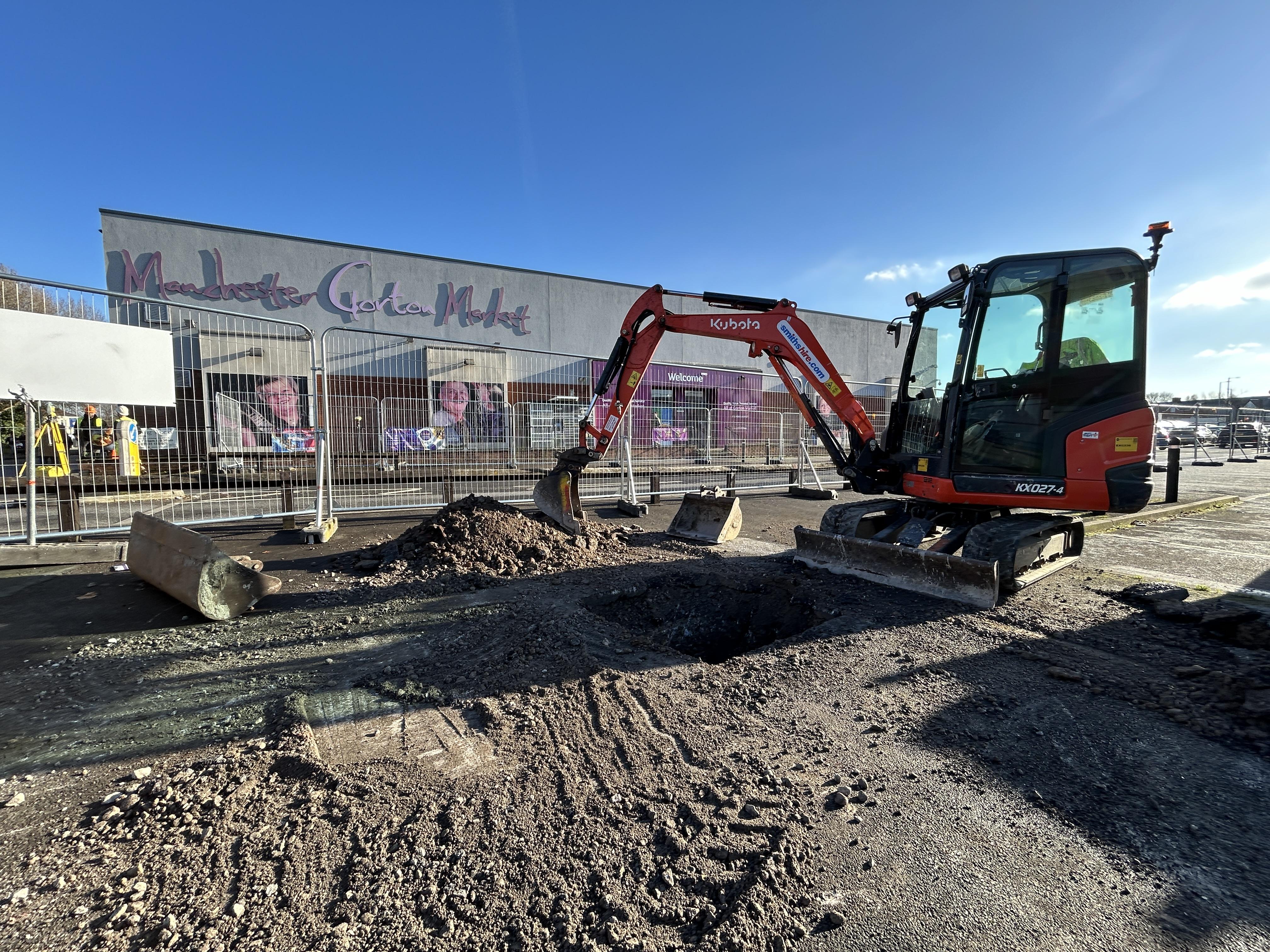 A digger poised to start work at Gorton town centre with Market in the background