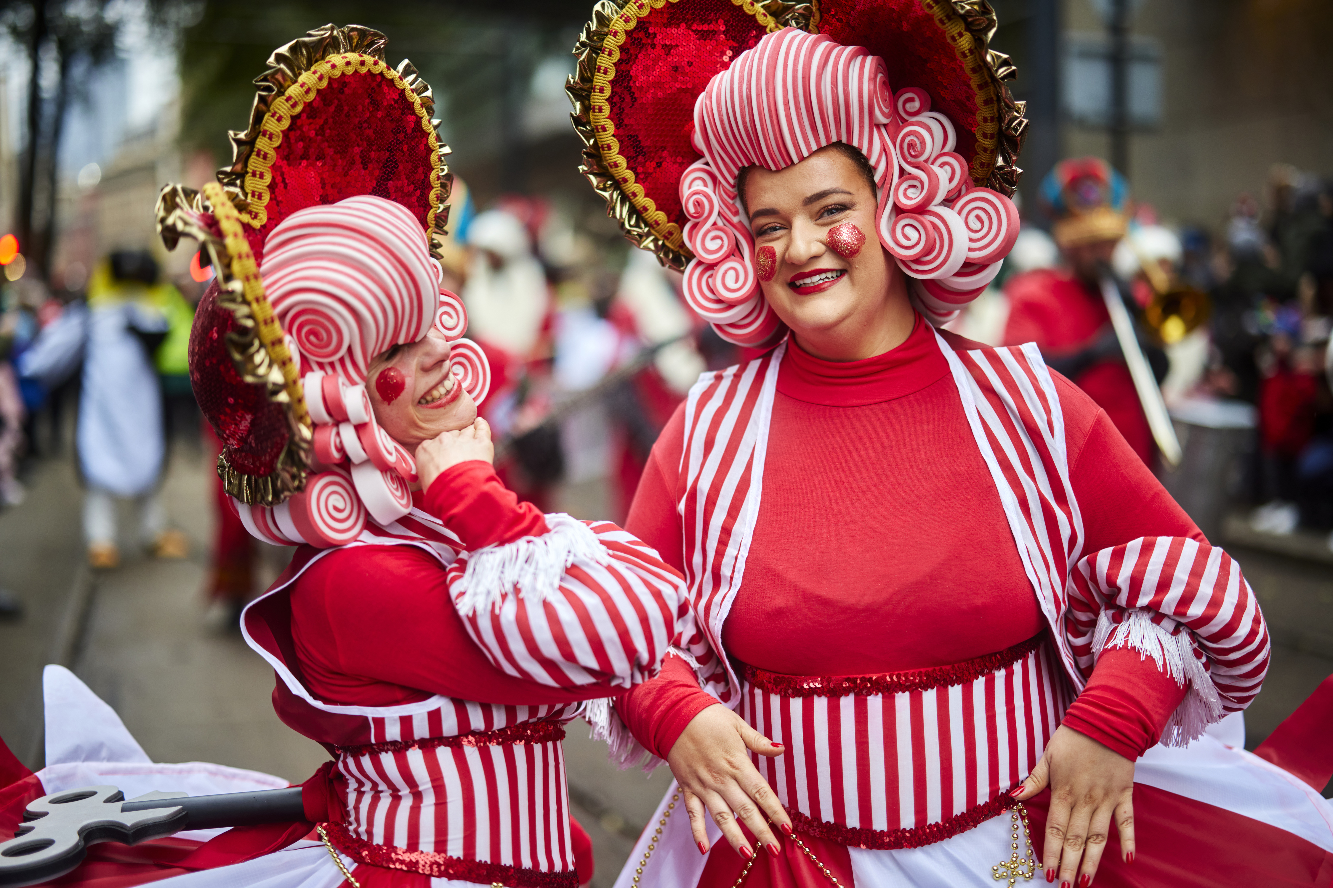 Street scene with crowds in the background. Two women dressed as larger than lift characters in bright red and white striped costumes and wearing big theatrical blonde wigs and hats, smiling as they perform to the camera