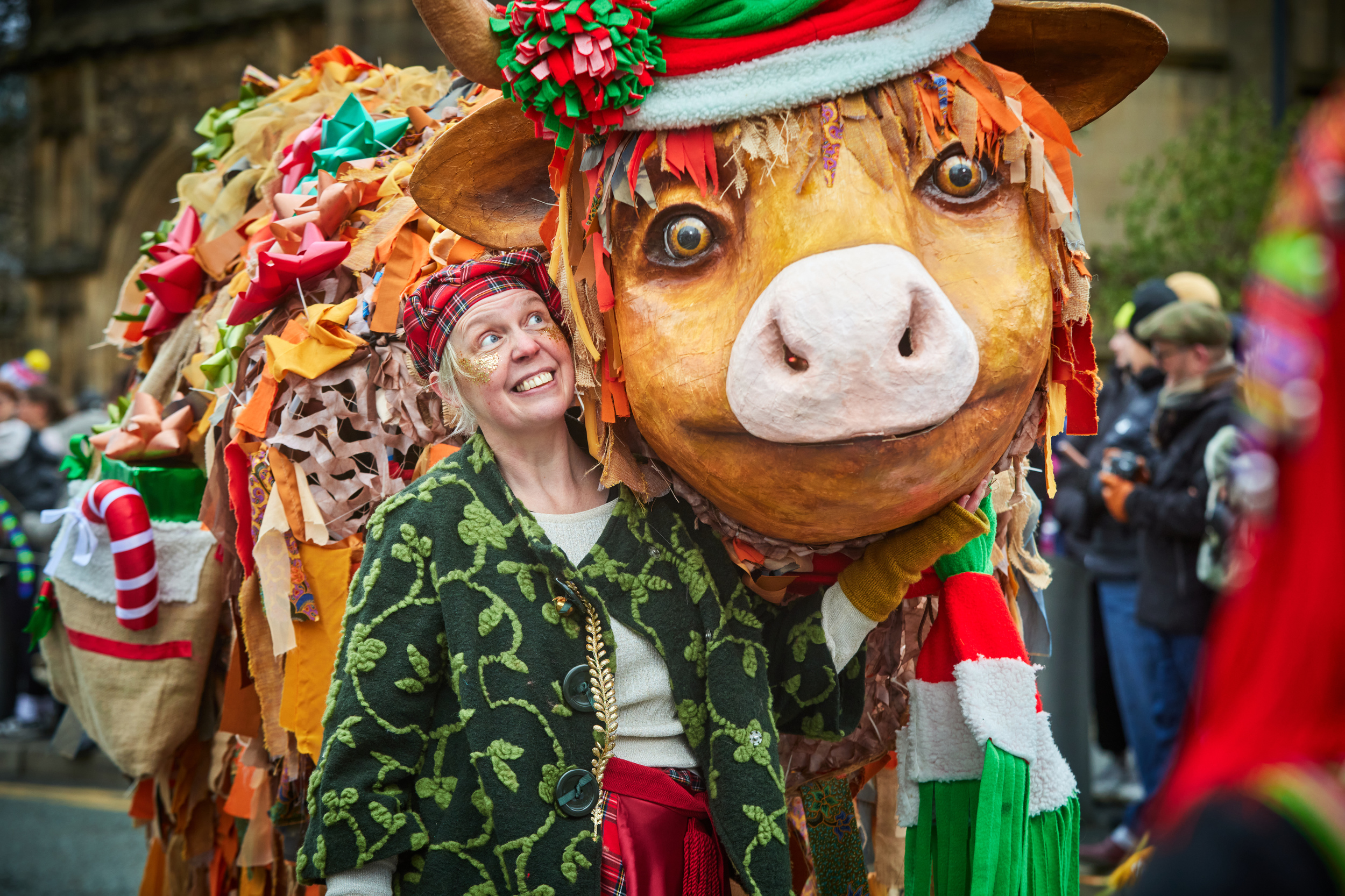 woman wearing a flamboyant outfit in Christmas colours smiling up at an oversize Christmas cow decorated with candy canes and tinsel.  They are in a parade