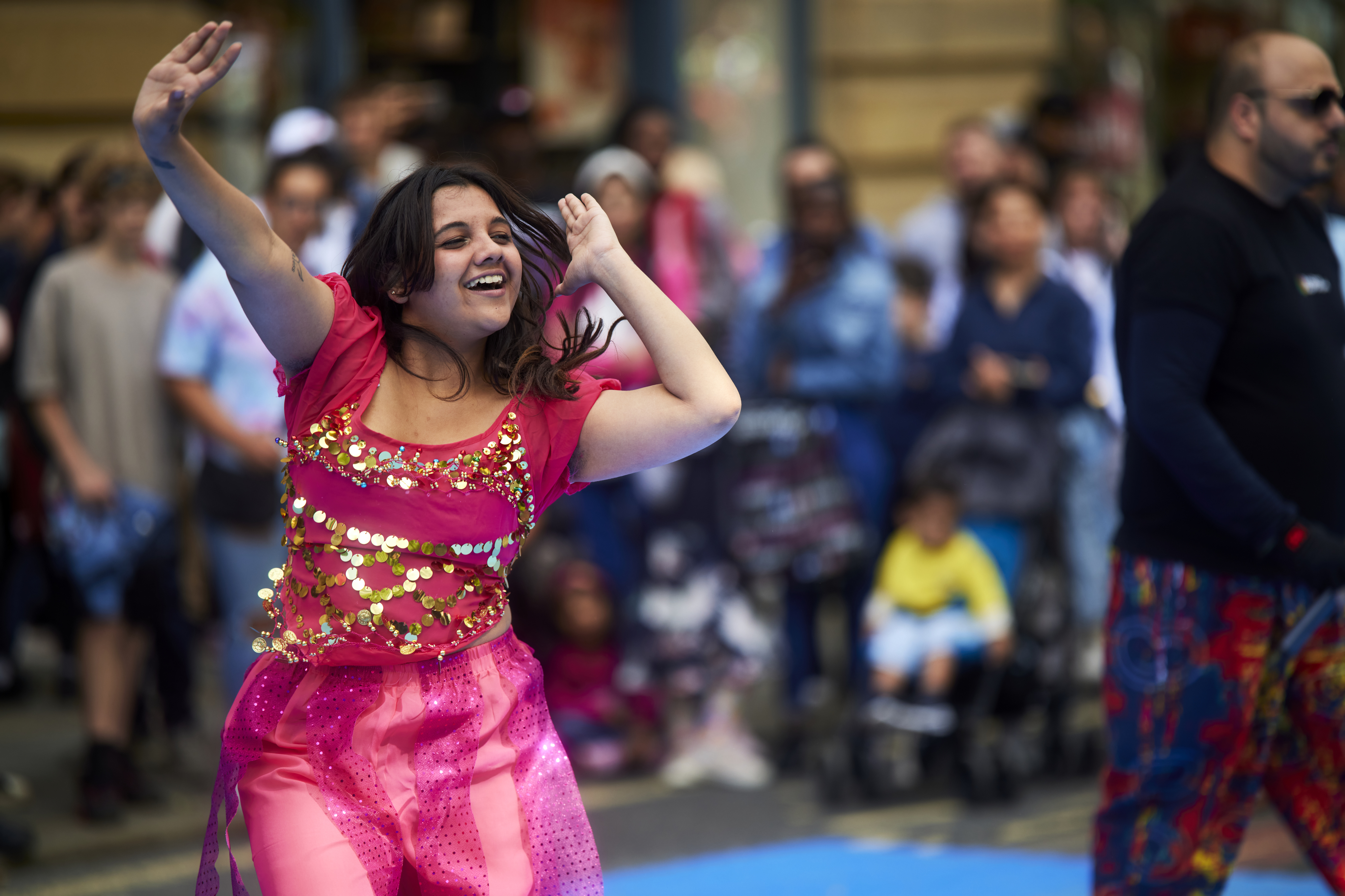 Street scene, young woman dressed in pink smiles as she dances with her arms in the air in front of crowds of people