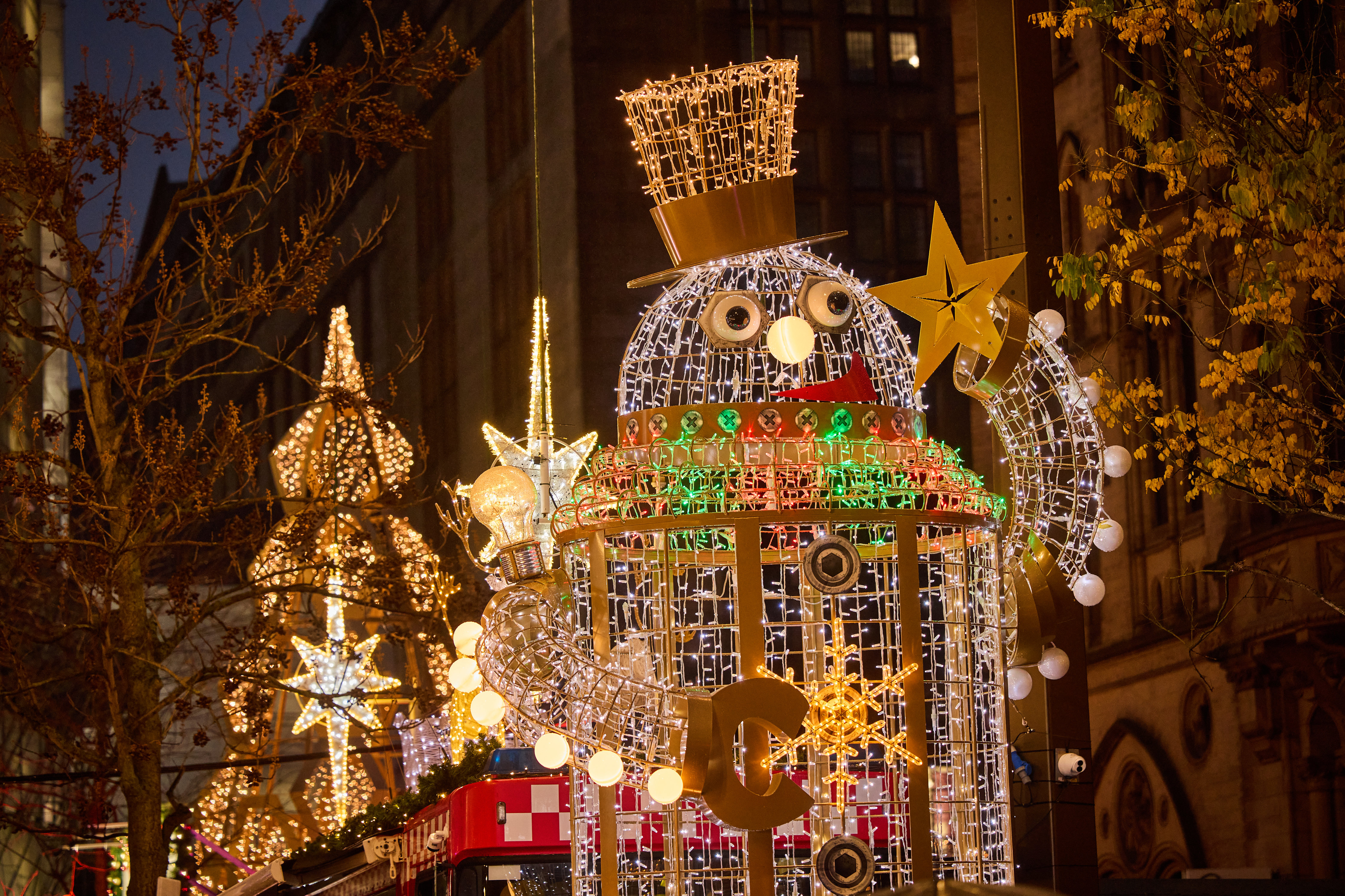 Street scene at night with a giant sized snowman figure wearing a top hat - all made out of twinkling golden fairy lights