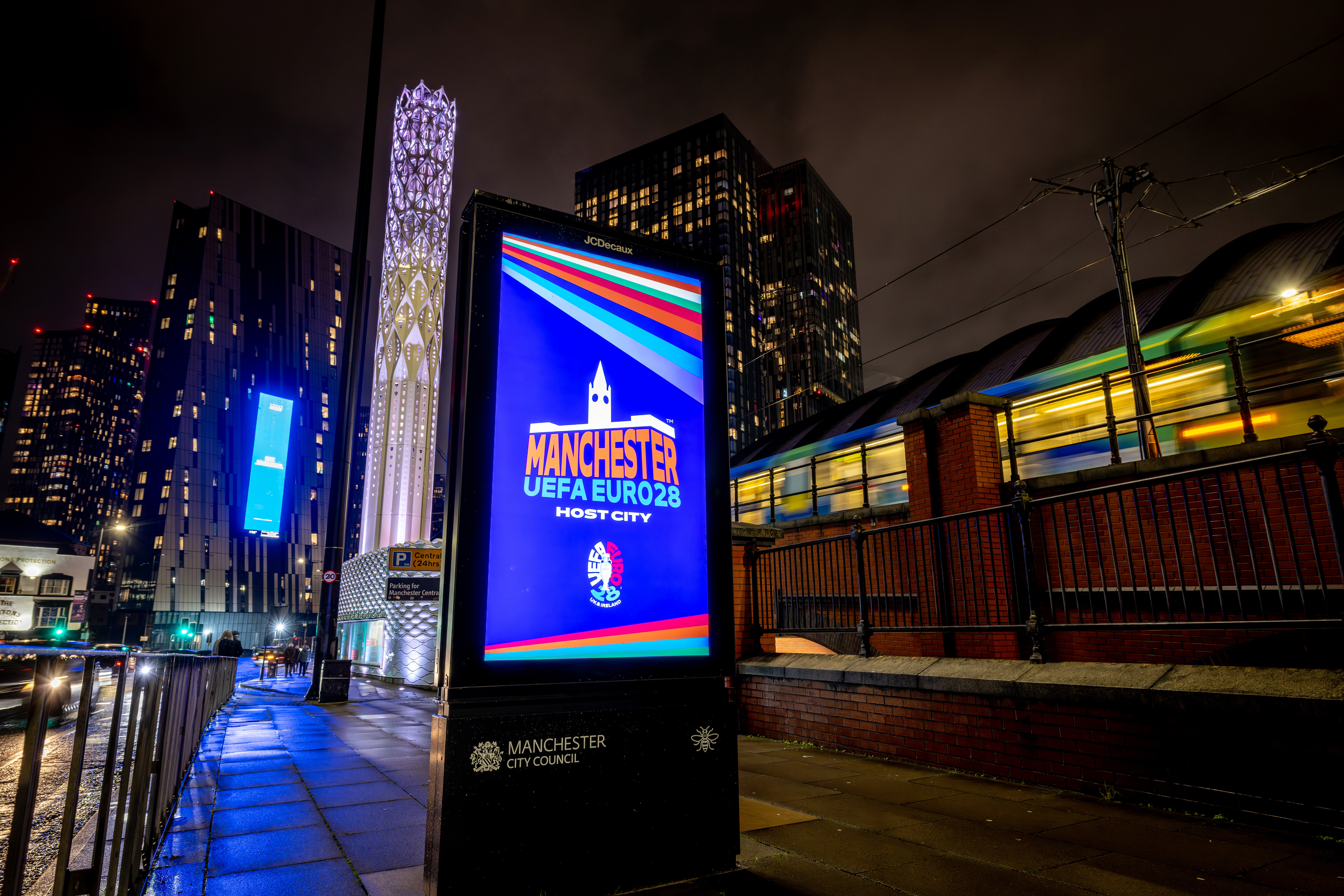 Street scene at night, an on-pavement digital advertising screen displays a bright blue logo bearing the words Manchester UEFA EURO 28 Host City on it, as a tram goes past to the right of the display board