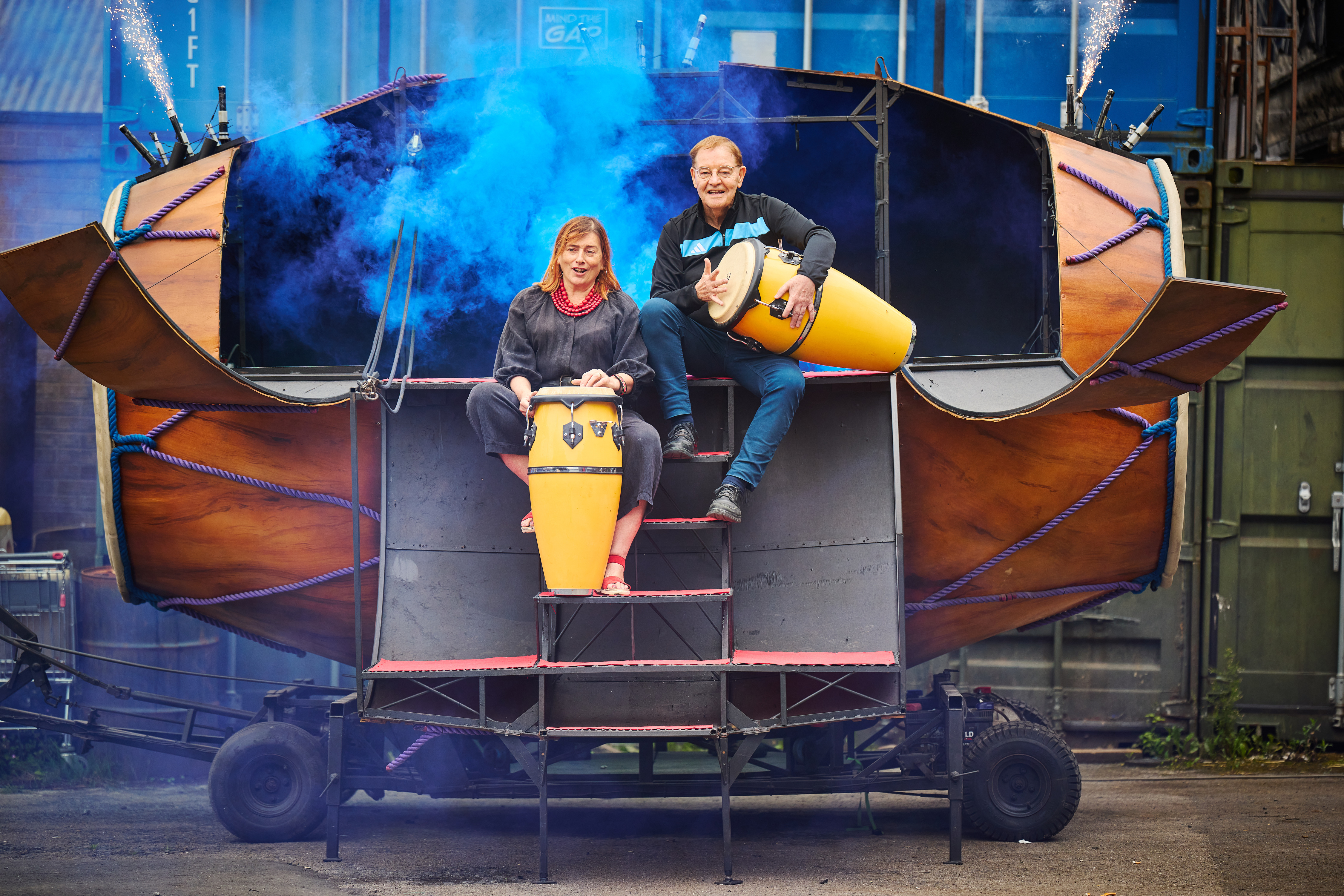 Two adults sitting on steps, each playing a big conga drum.  Behind them is blue smoke and fireworks going off.