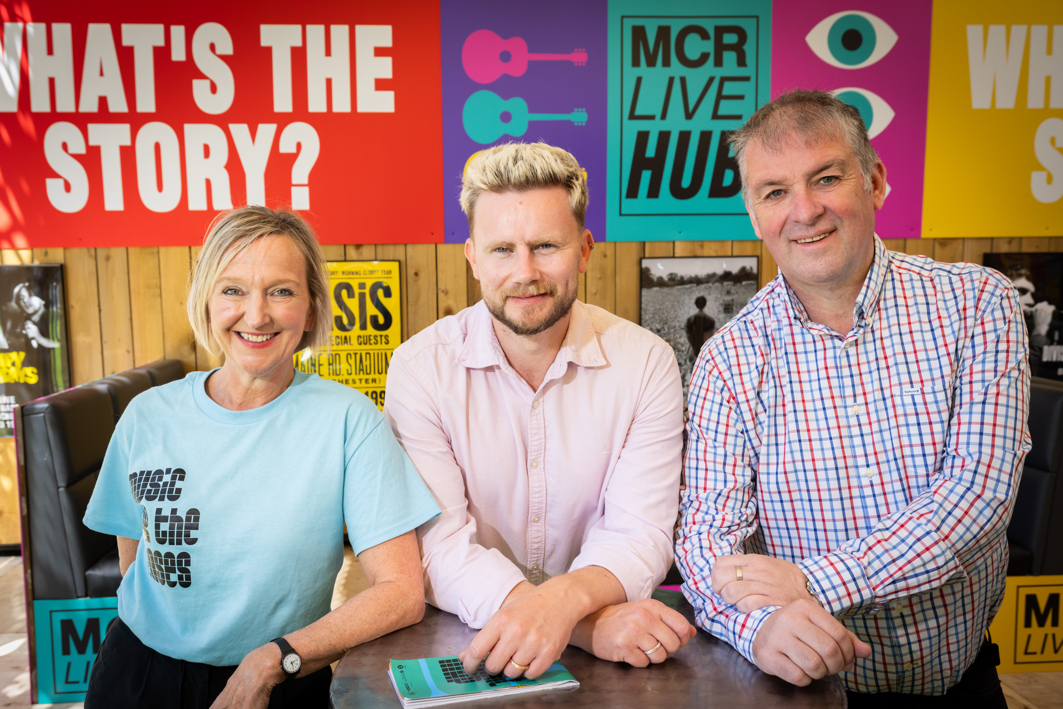 Three people smiling looking at the camera, brightly coloured posters on a wall behind them
