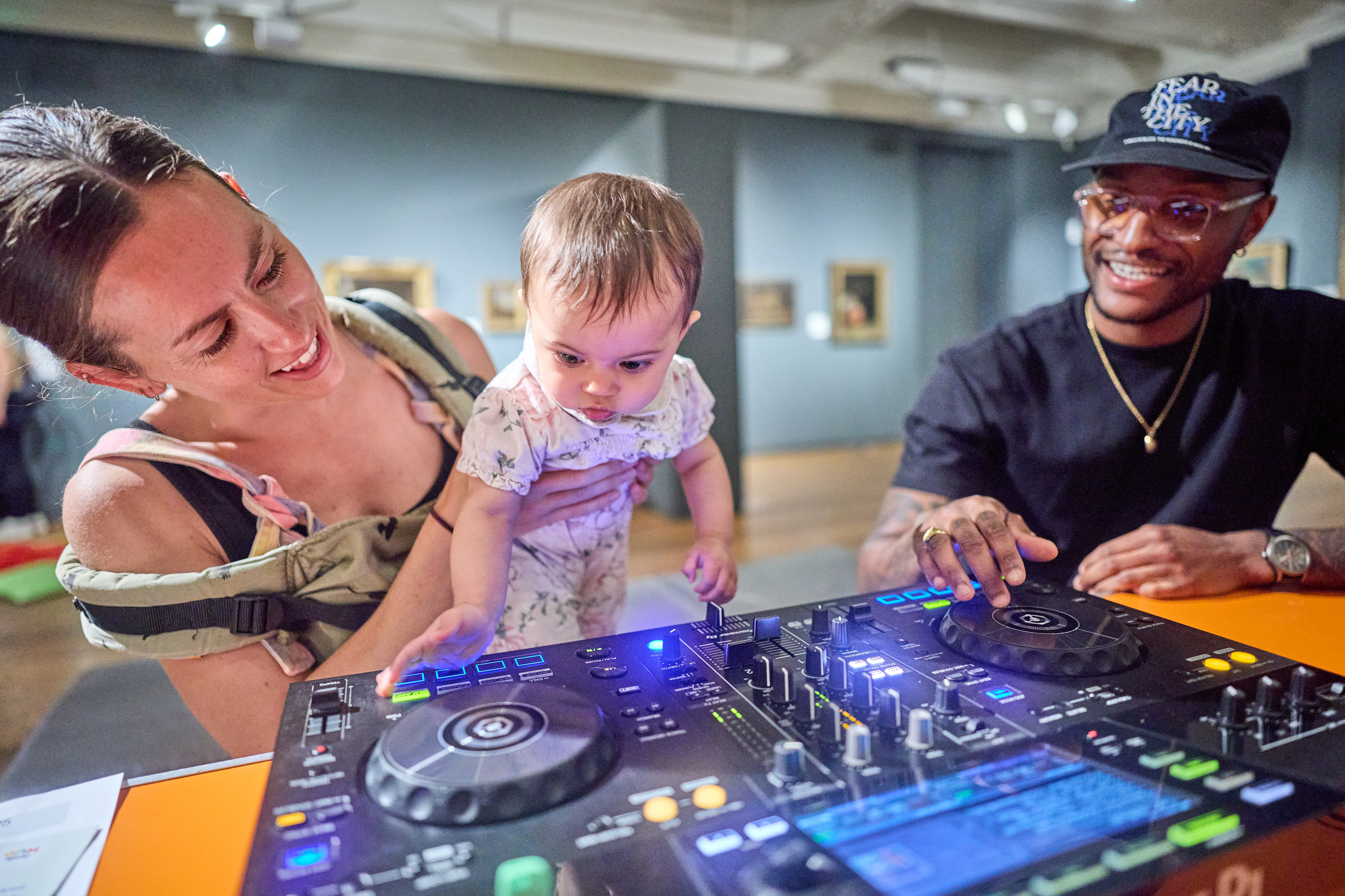 Woman holding a baby who is reaching out to touch DJ decks. Man watching and smiling next to them.