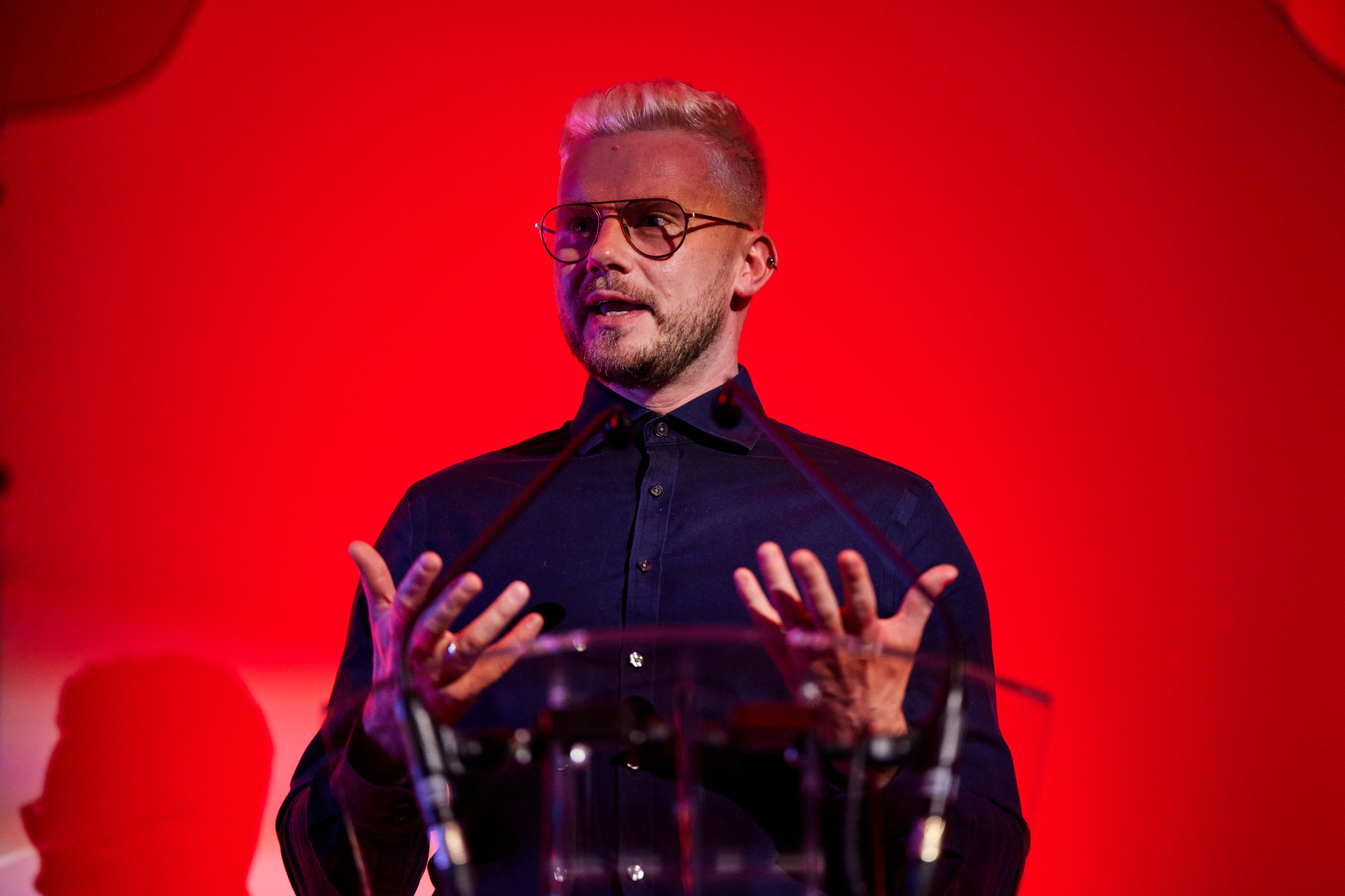 Man, short hair, wearing a dark coloured shirt standing behind microphones speaking animatedly to an unseen audience