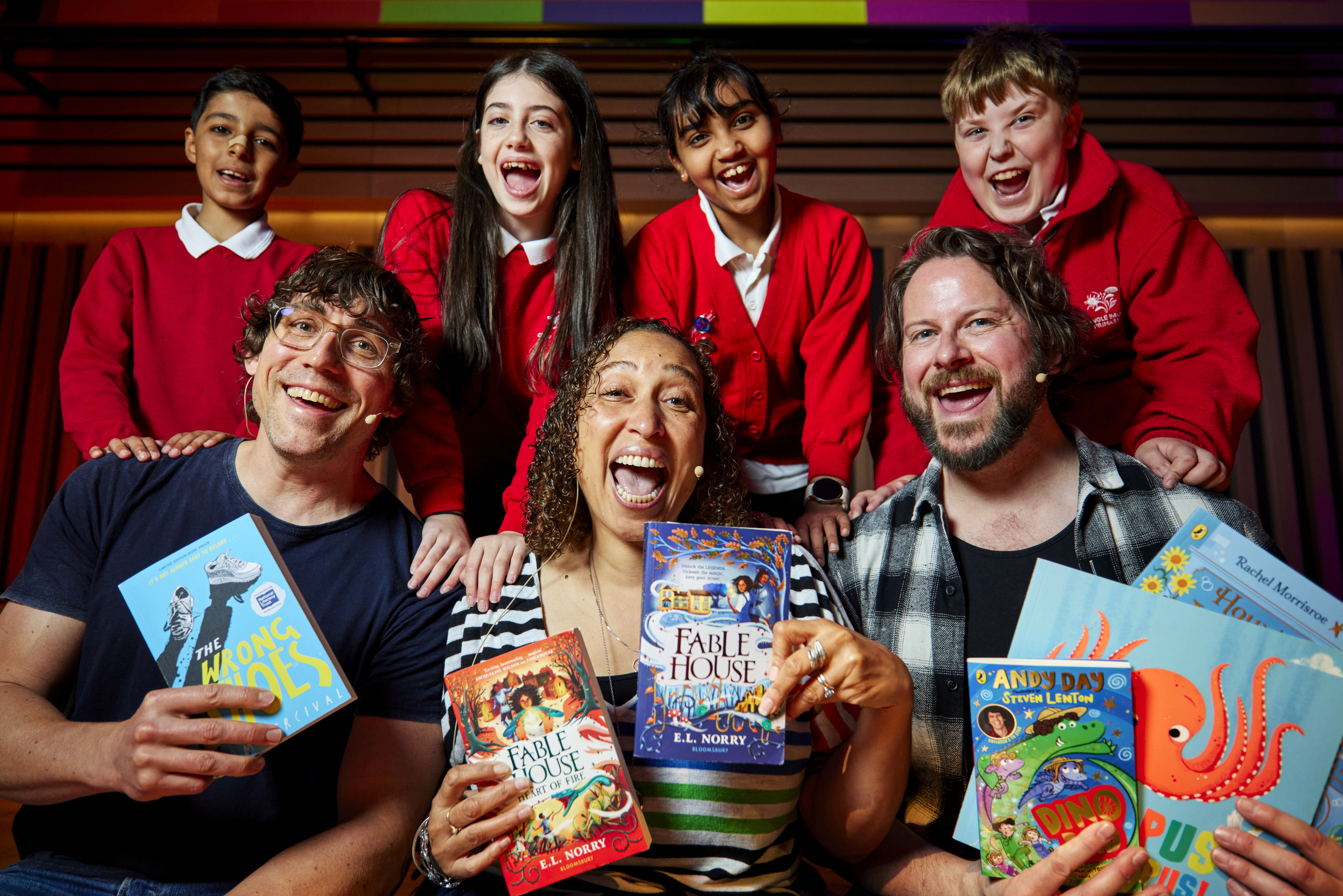 Group of three adults sitting at the front of the group holding books up in their hands, with four primary school aged children wearing red uniforms standing behind them - all smiling