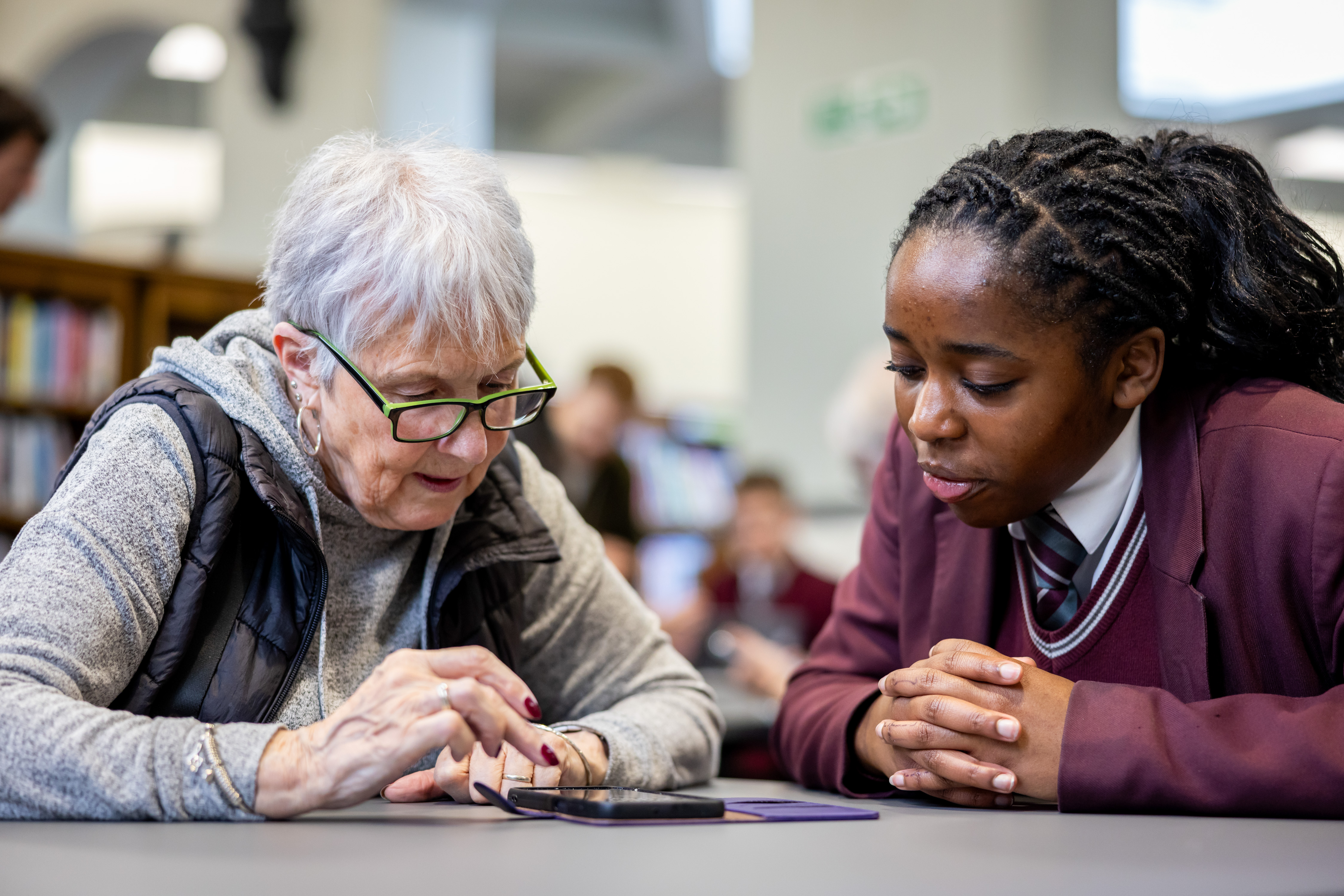 An older woman and teenage female school pupil sit together looking at a mobile phone