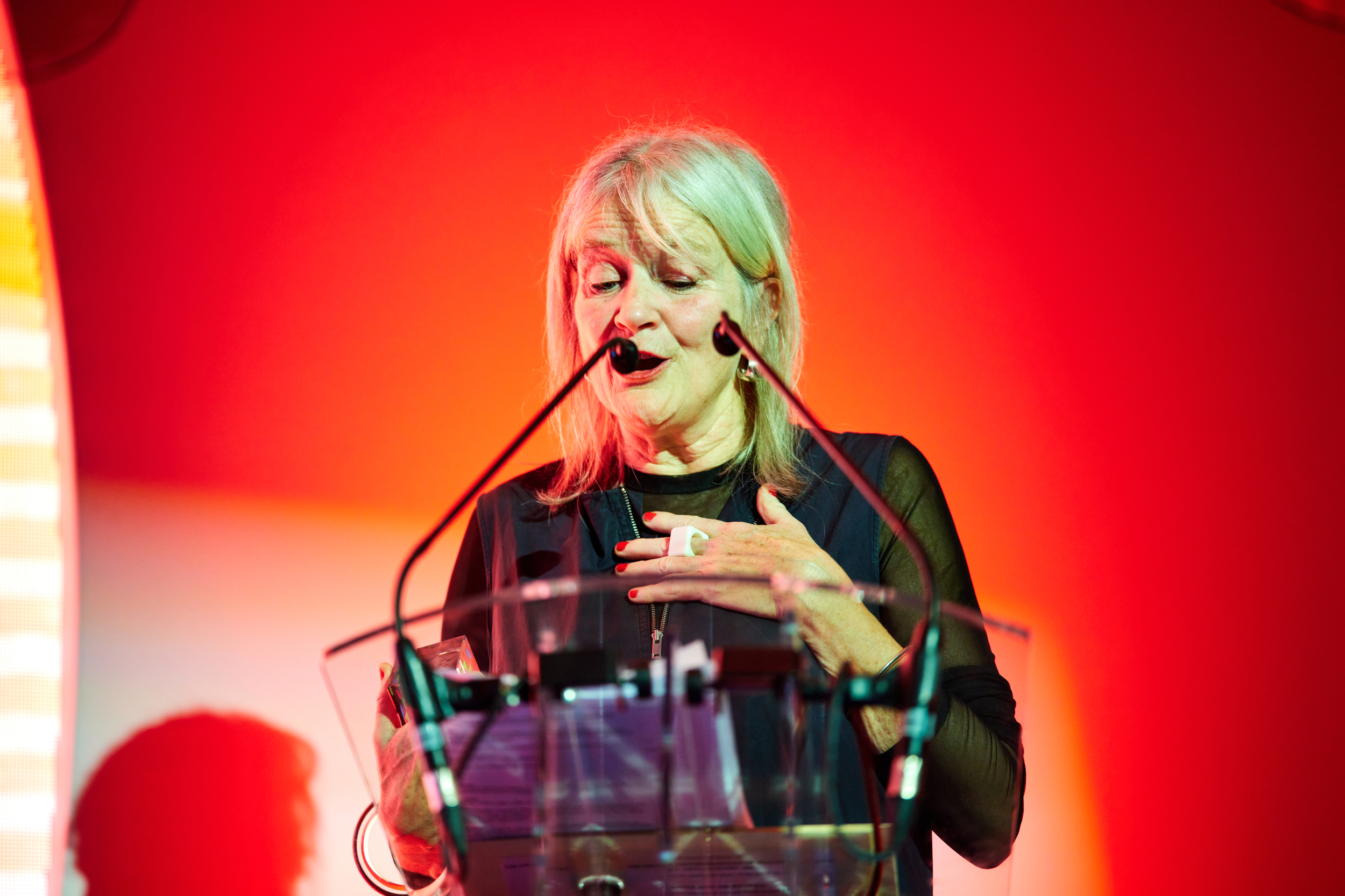 Woman, shoulder length hair, wearing a dark coloured dress, standing behind microphones speaking animatedly into an unseen audience
