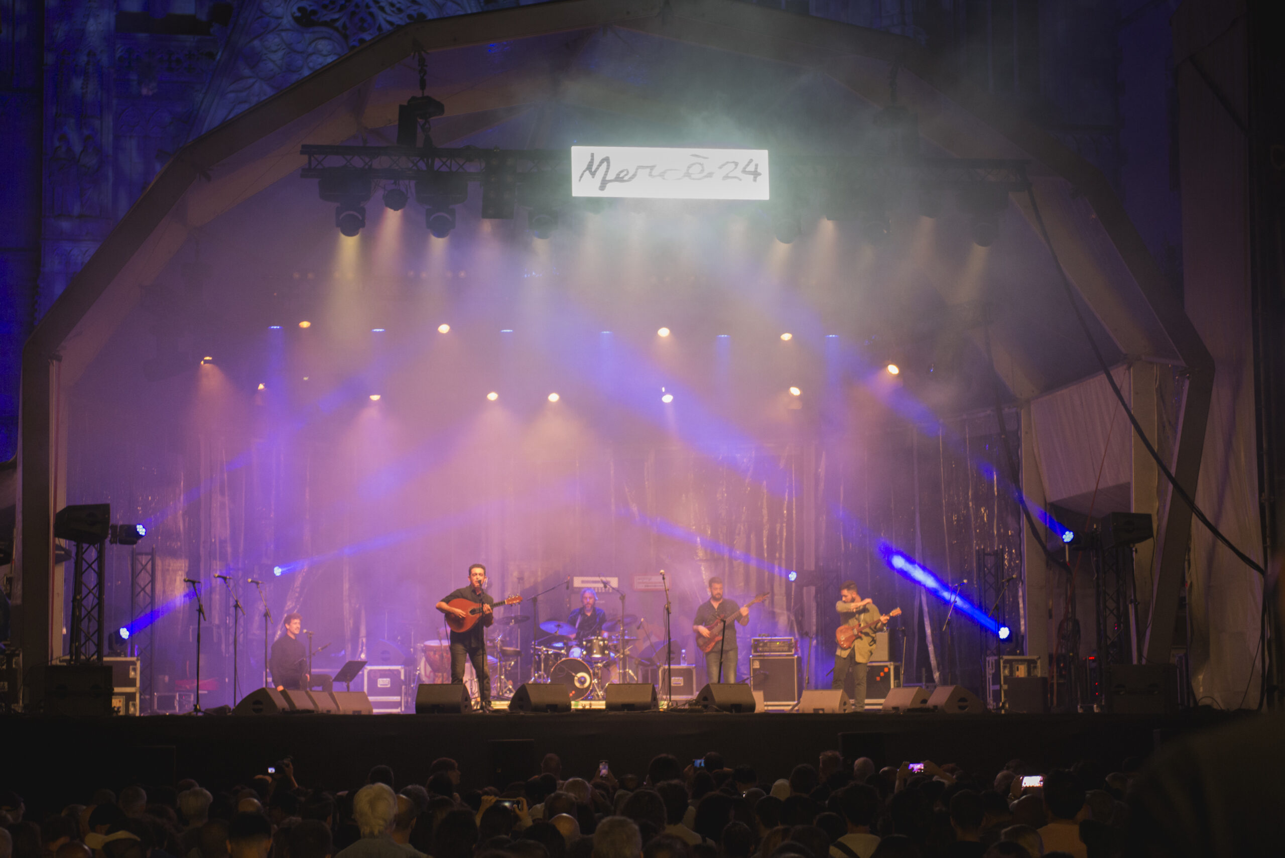 Night time view of a big outdoor stage, pink and orange lights shining on musicians playing on the stage Night time view of a big outdoor stage, pink and orange lights shining on musicians playing on the stage