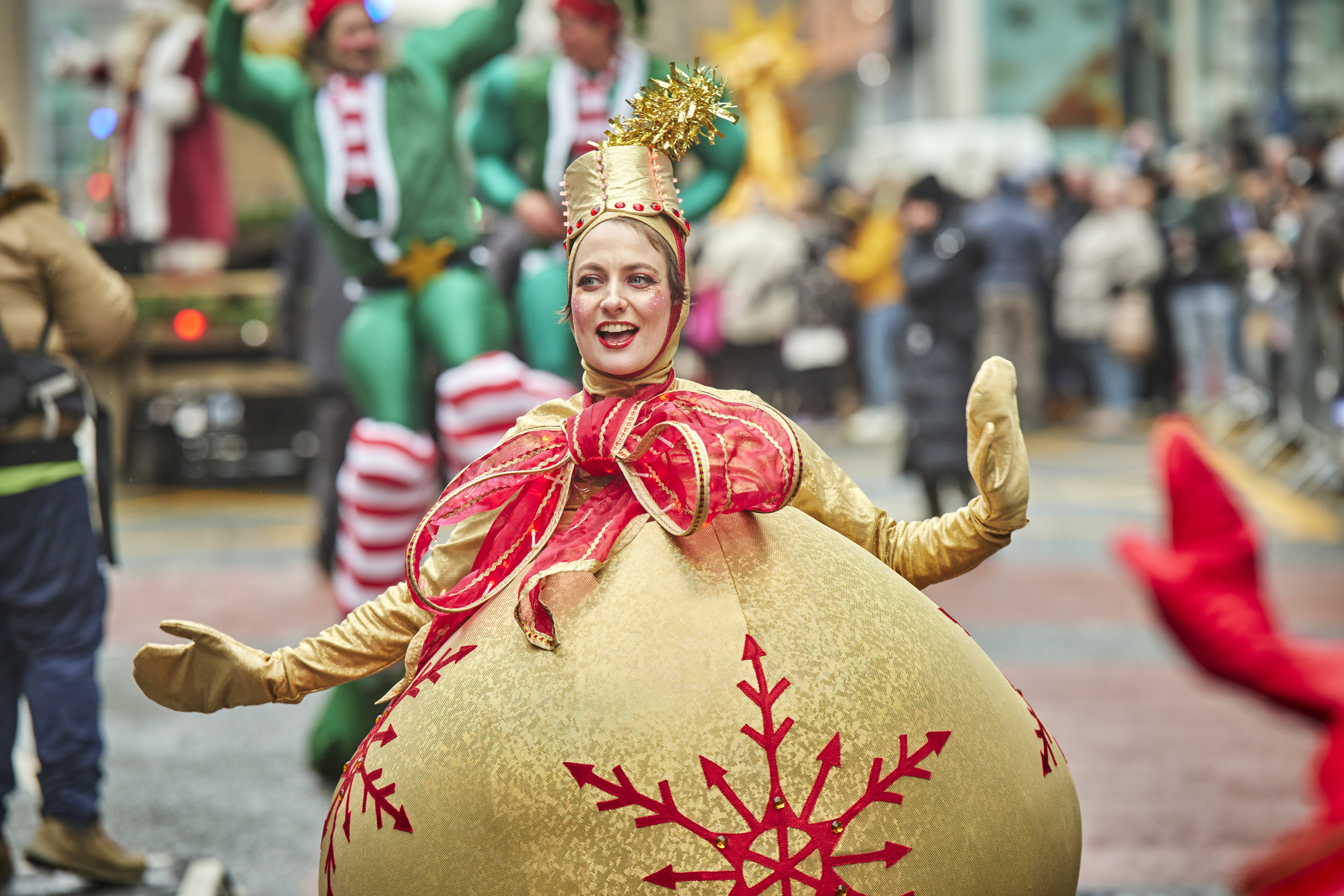 Street scene showing a woman dressed as a giant golden Christmas bauble with a red ribbon on top and crowds in the background