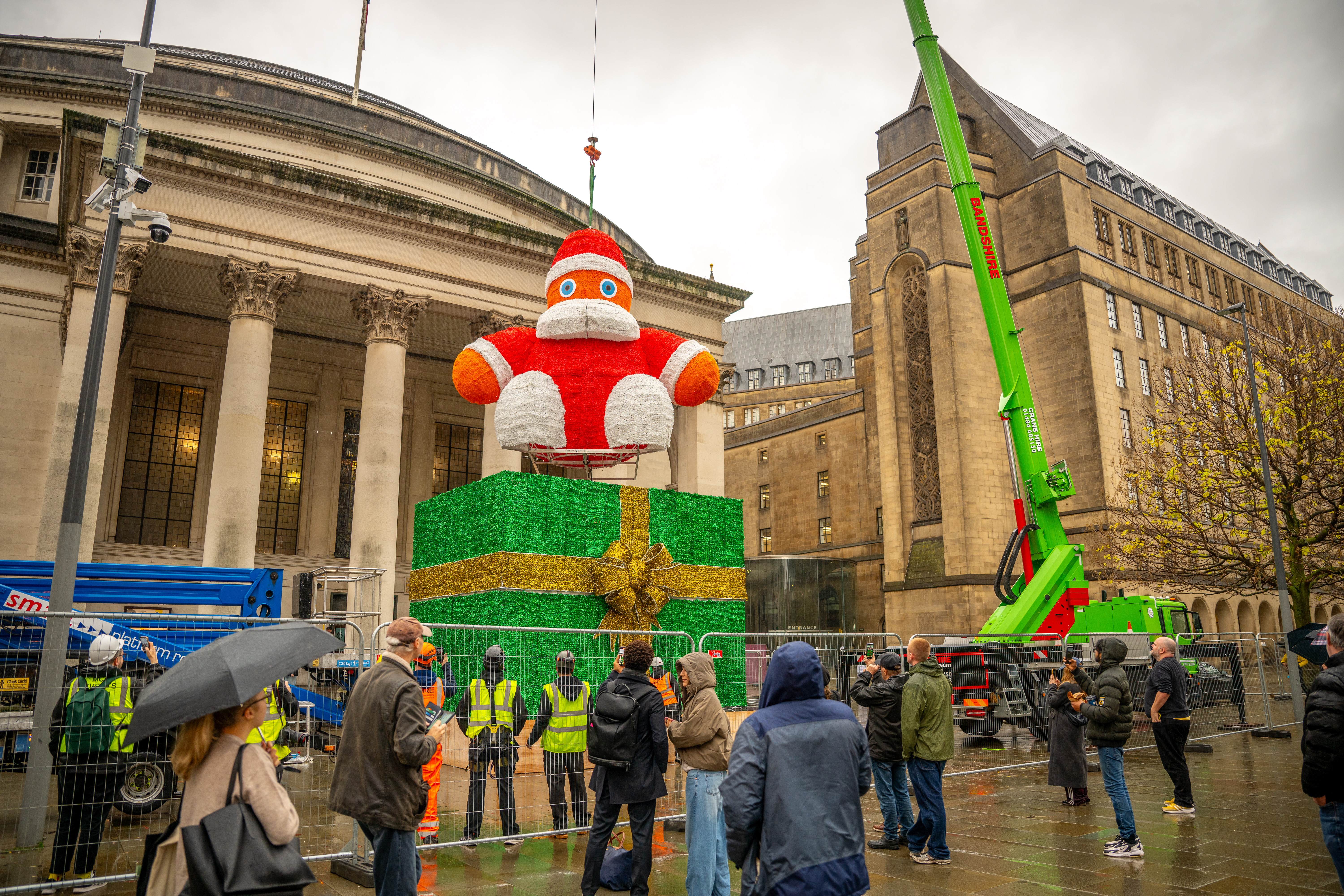 A giant Santa model is being lifted high in the air by a crane onto a bright green base shaped like a christmas present, all watched by people standing on the ground and looking up