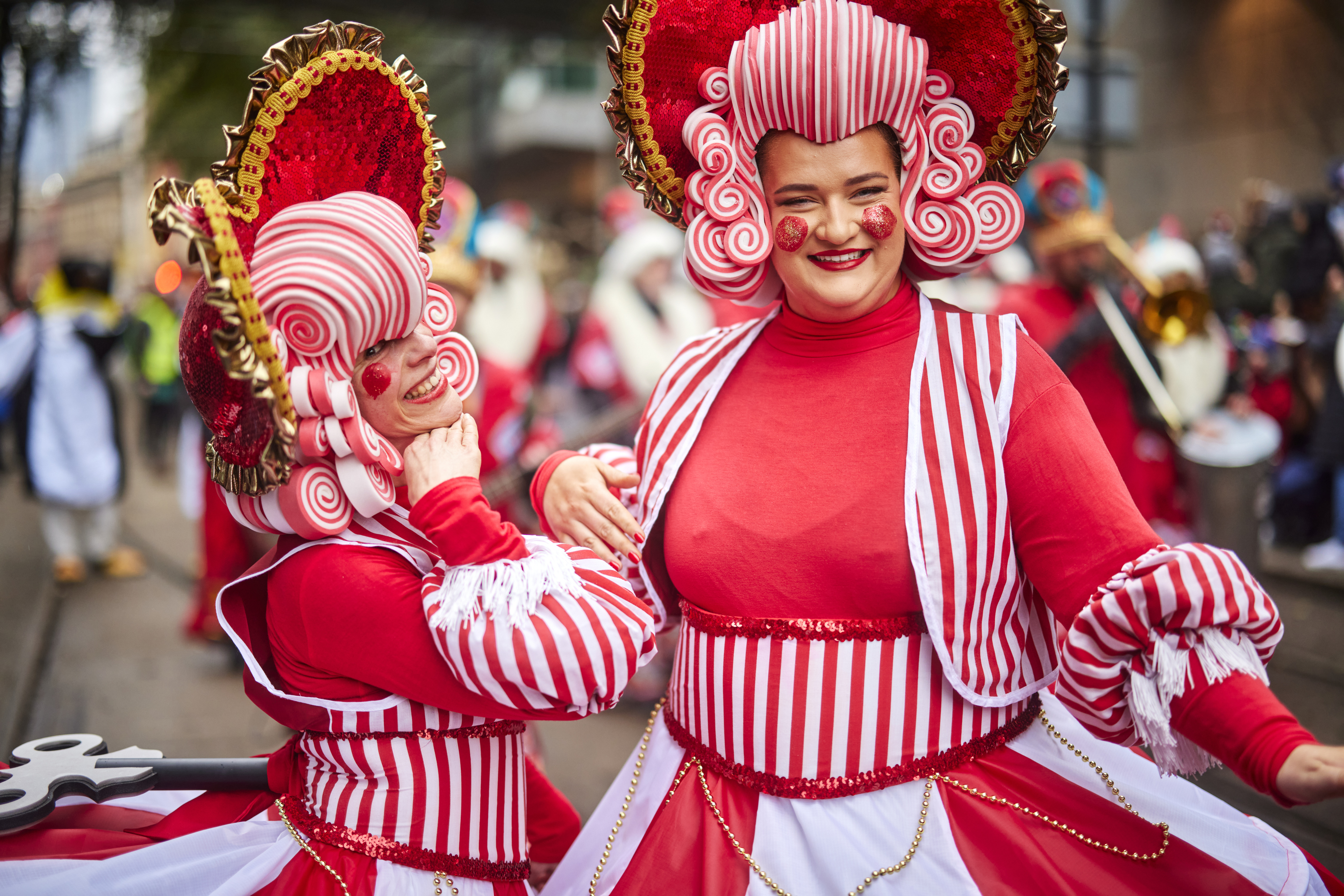 Sreet scene, a parade. Two smiling people play-acting, dressed as larger than life characters wearing red costumes with white stripes and big wigs