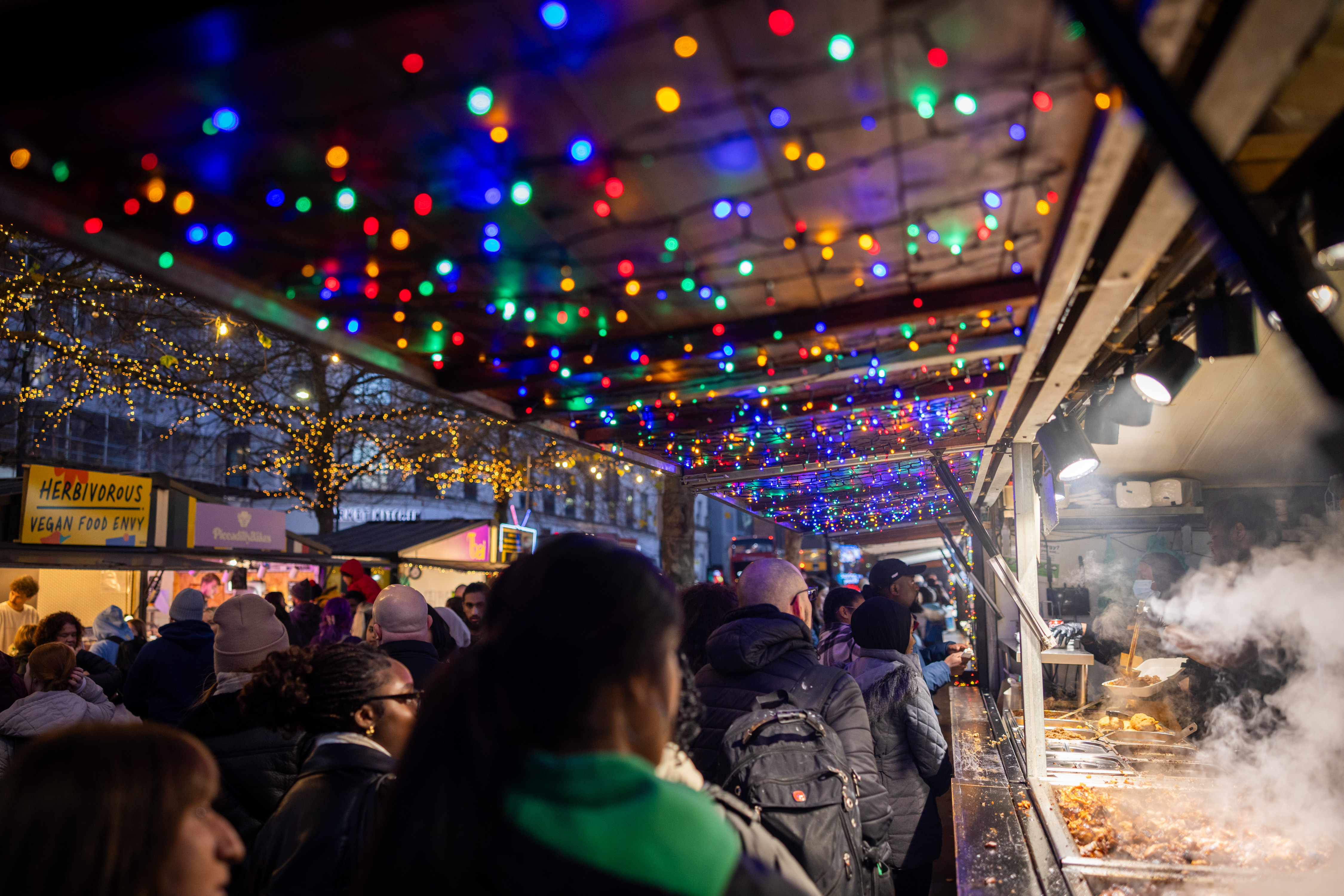 Night time street scene. Crowds of people walking past festive stalls, some with food cooking and steam rising from pans. Twinkling lights overhead. 