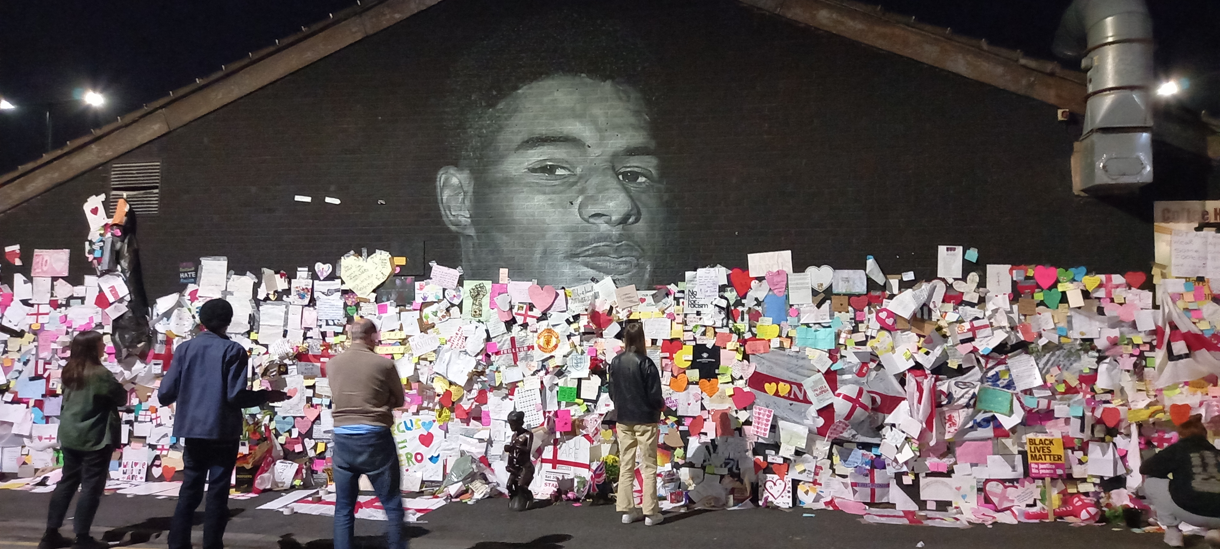 A black wall with a mural of the face of footballer Marcus Rashford on it, the wall is covered in thousands of pieces of paper in all colours, shapes and sizes