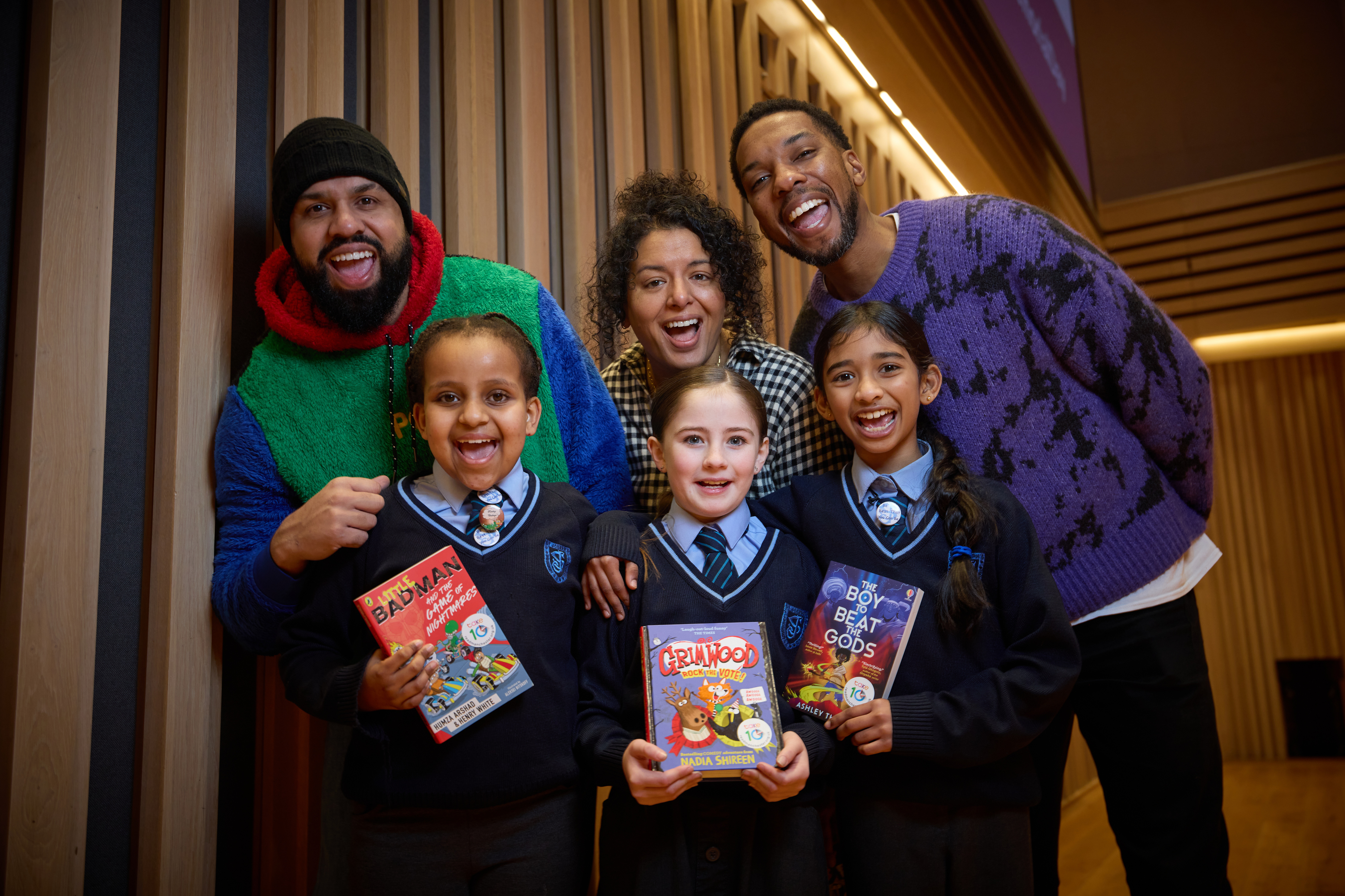 4 children holding books and smiling at the camera with 3 grown-ups behind them also smiling