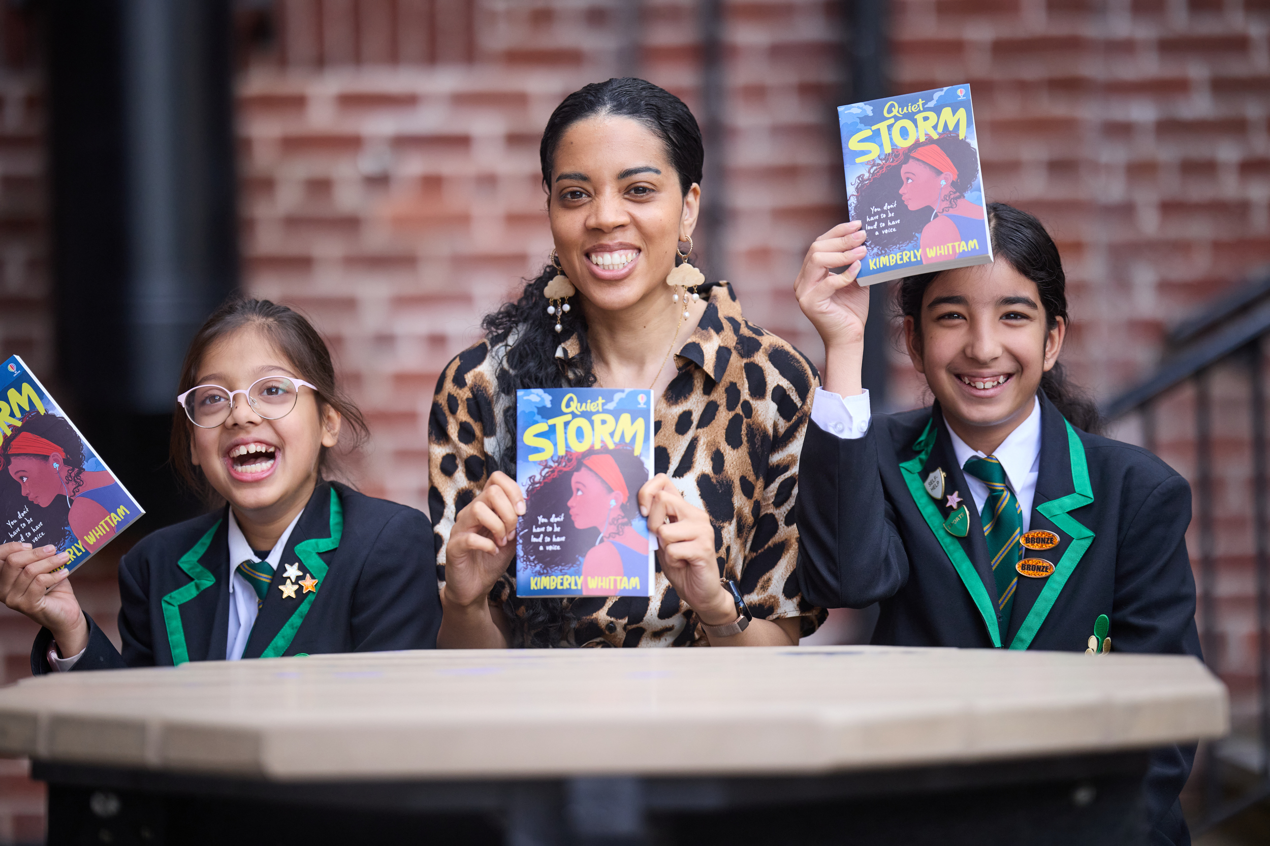 A woman sits inbetween two young school pupils wearing black blazers with green edging. They are each holding a copy of the same book and are all smiling.