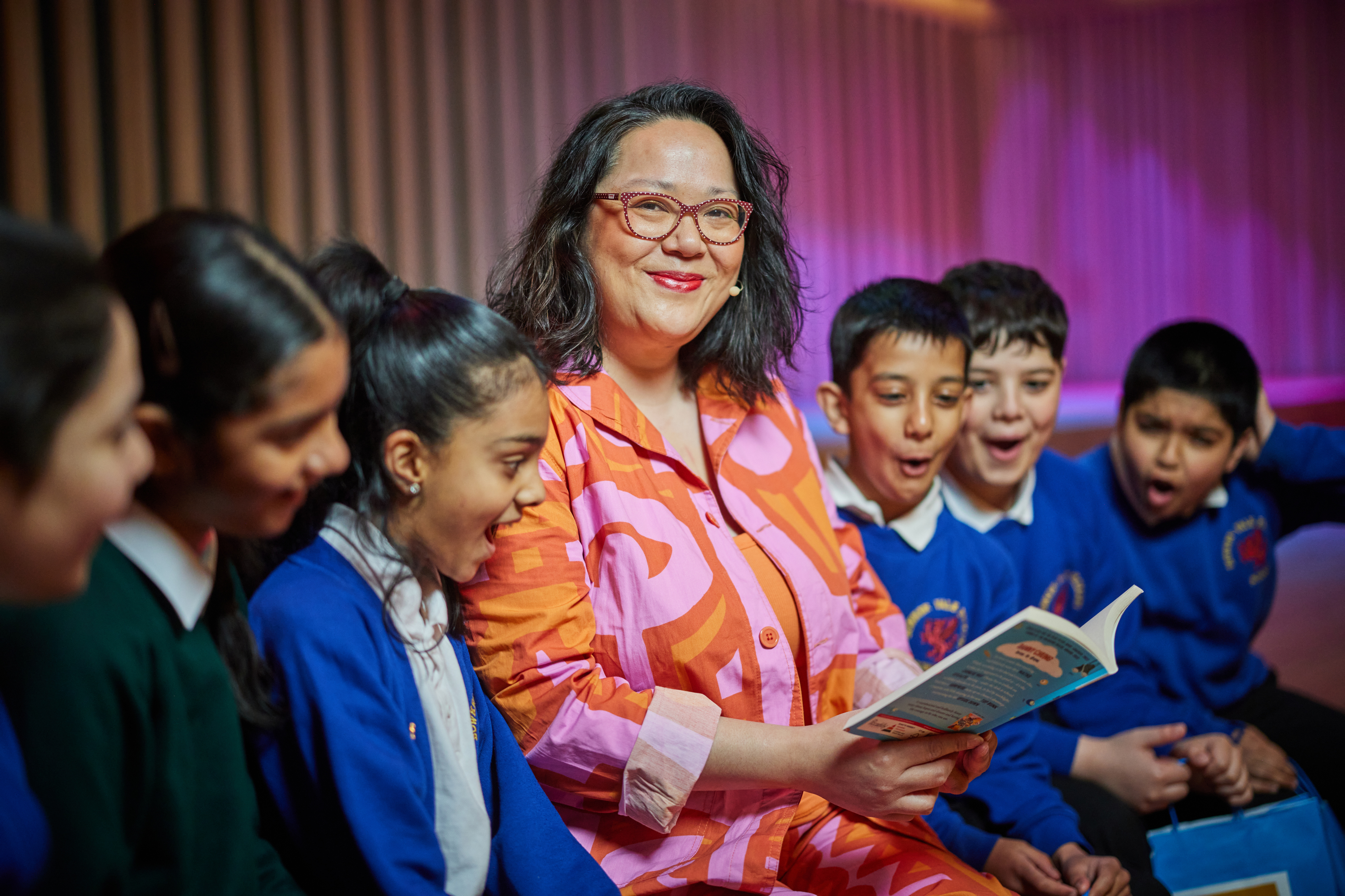 Woman sitting, holding an open book looking at the camera, surrounded either side by primary aged children looking at the book she is holding.