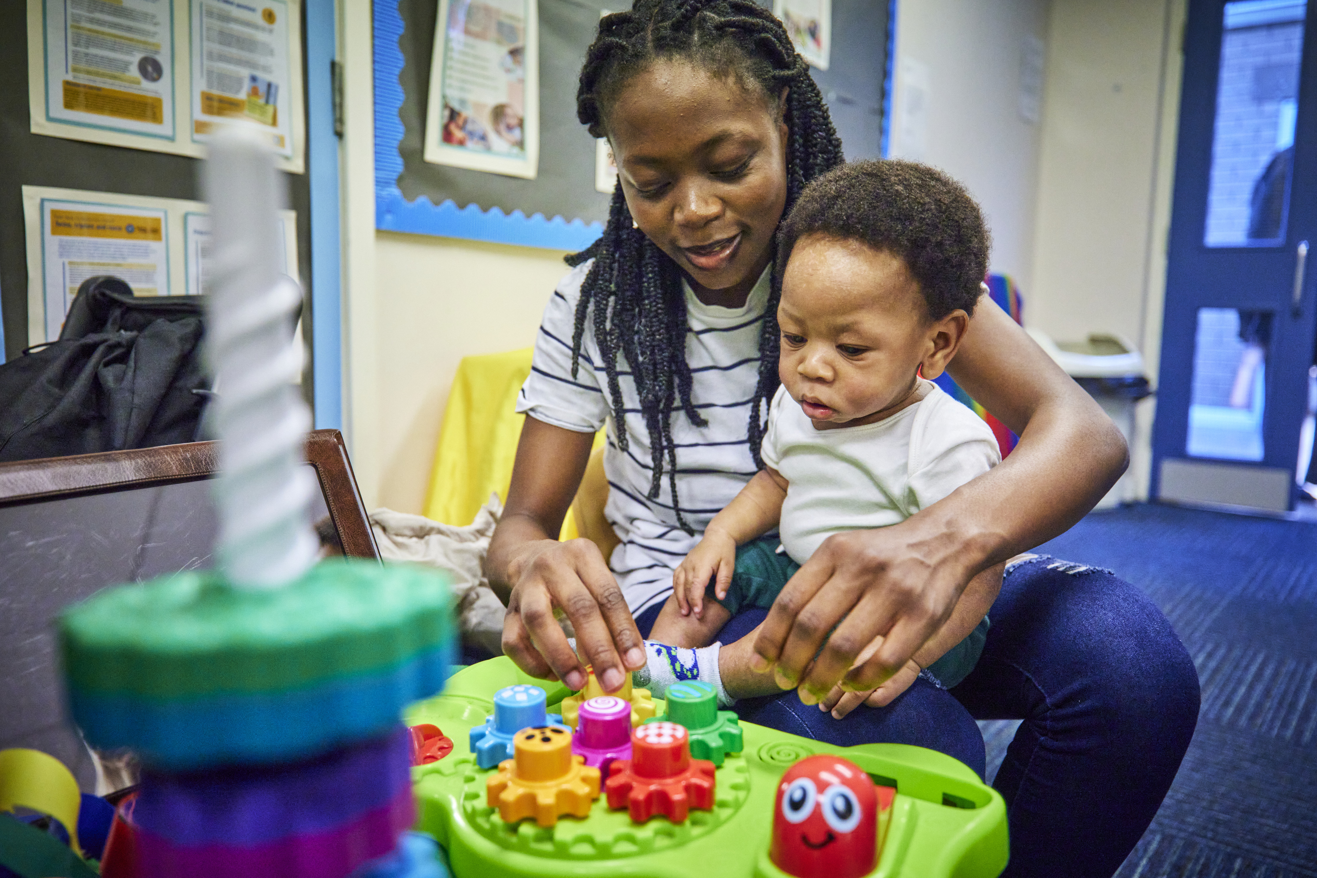 Woman smiling as she kneels with a small child on her lap, both playing with a brightly coloured toy