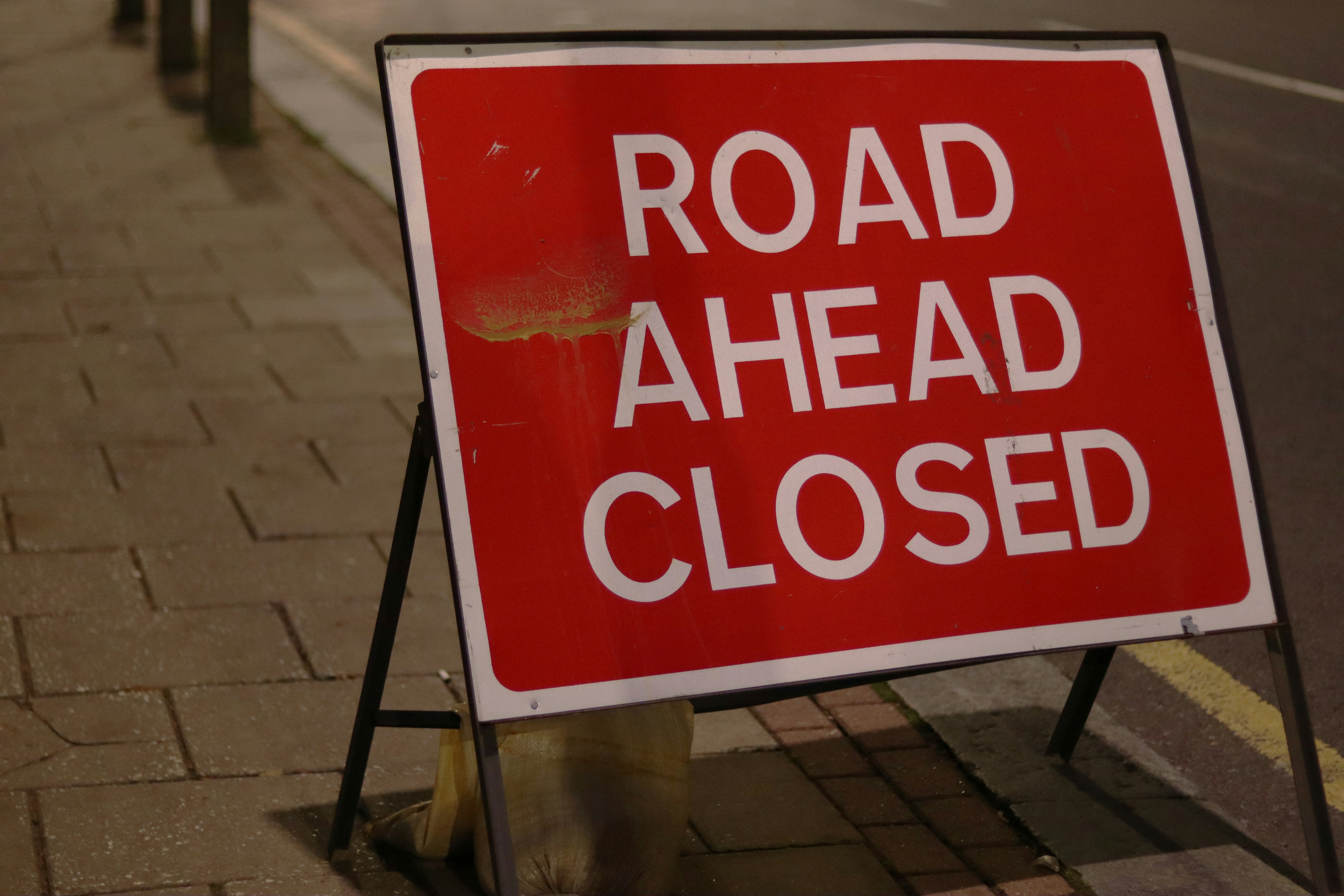 Red traffic sign on a pavement, which reads 'road ahead closed'.
