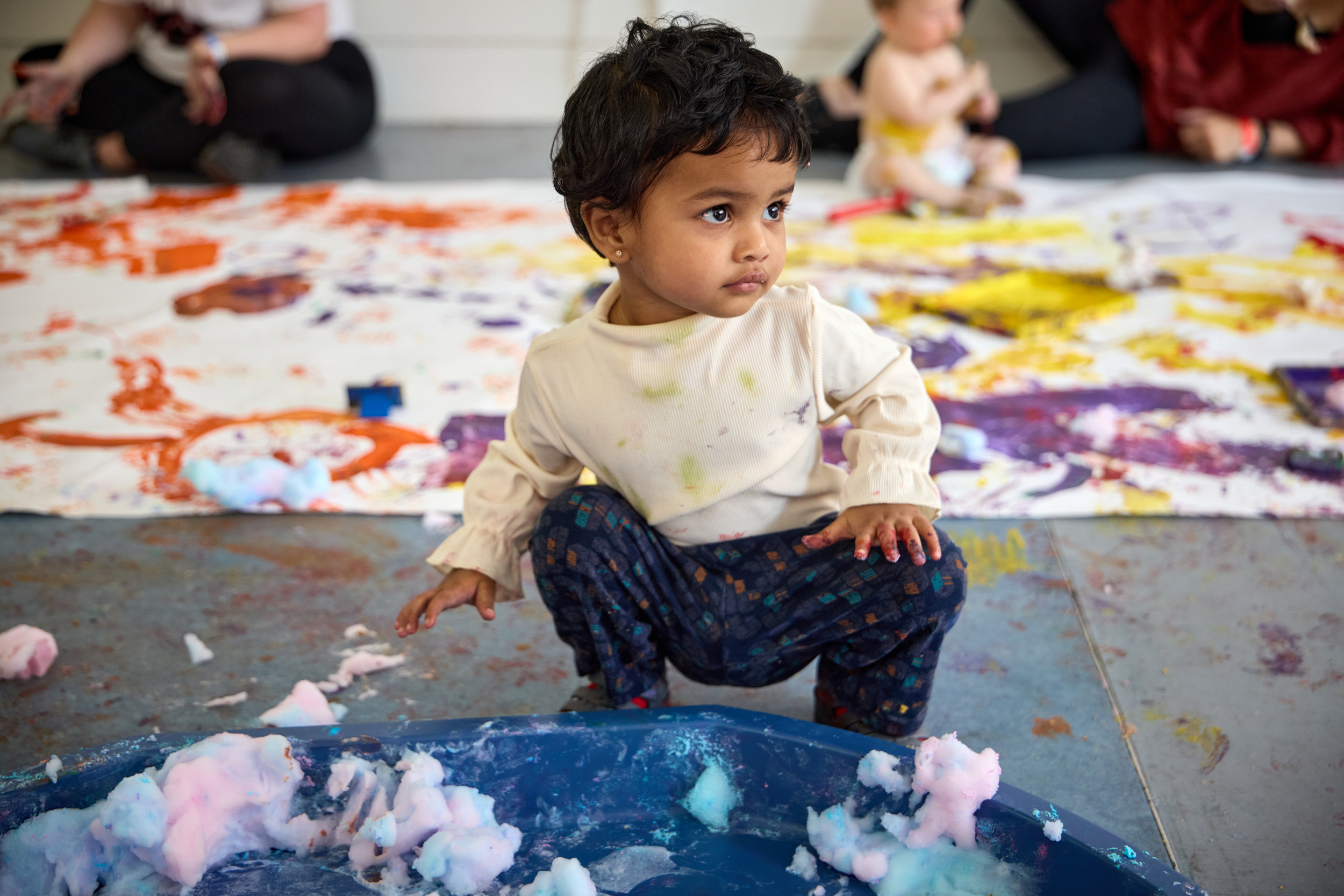 A toddler crouches on the floor behind her lots of white paper filled with brightly coloured splashes of paint