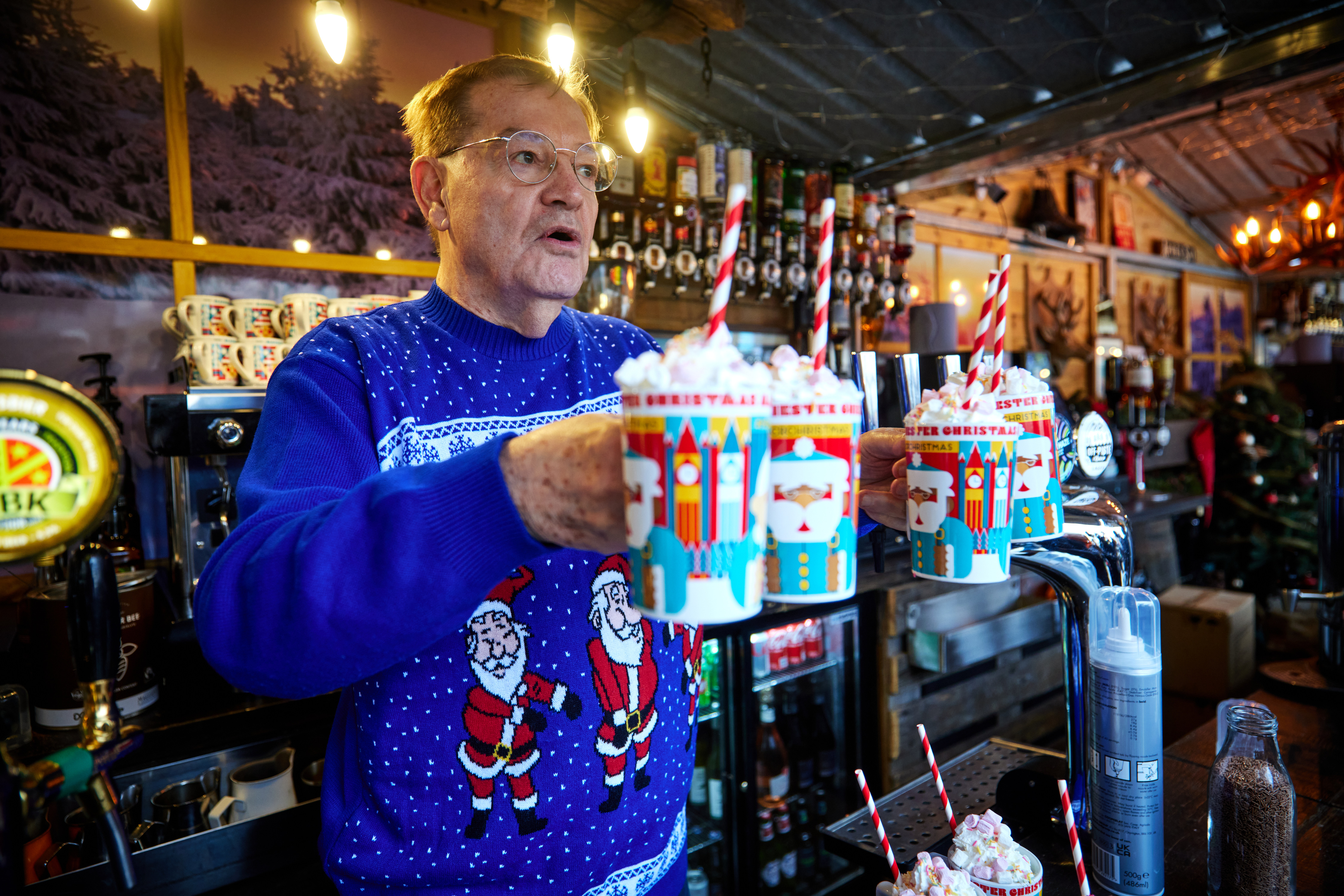Man wearing a blue Christmas jumper standing behind a bar holding 4 Christmas mugs each overflowing with whipped cream and marshmallows