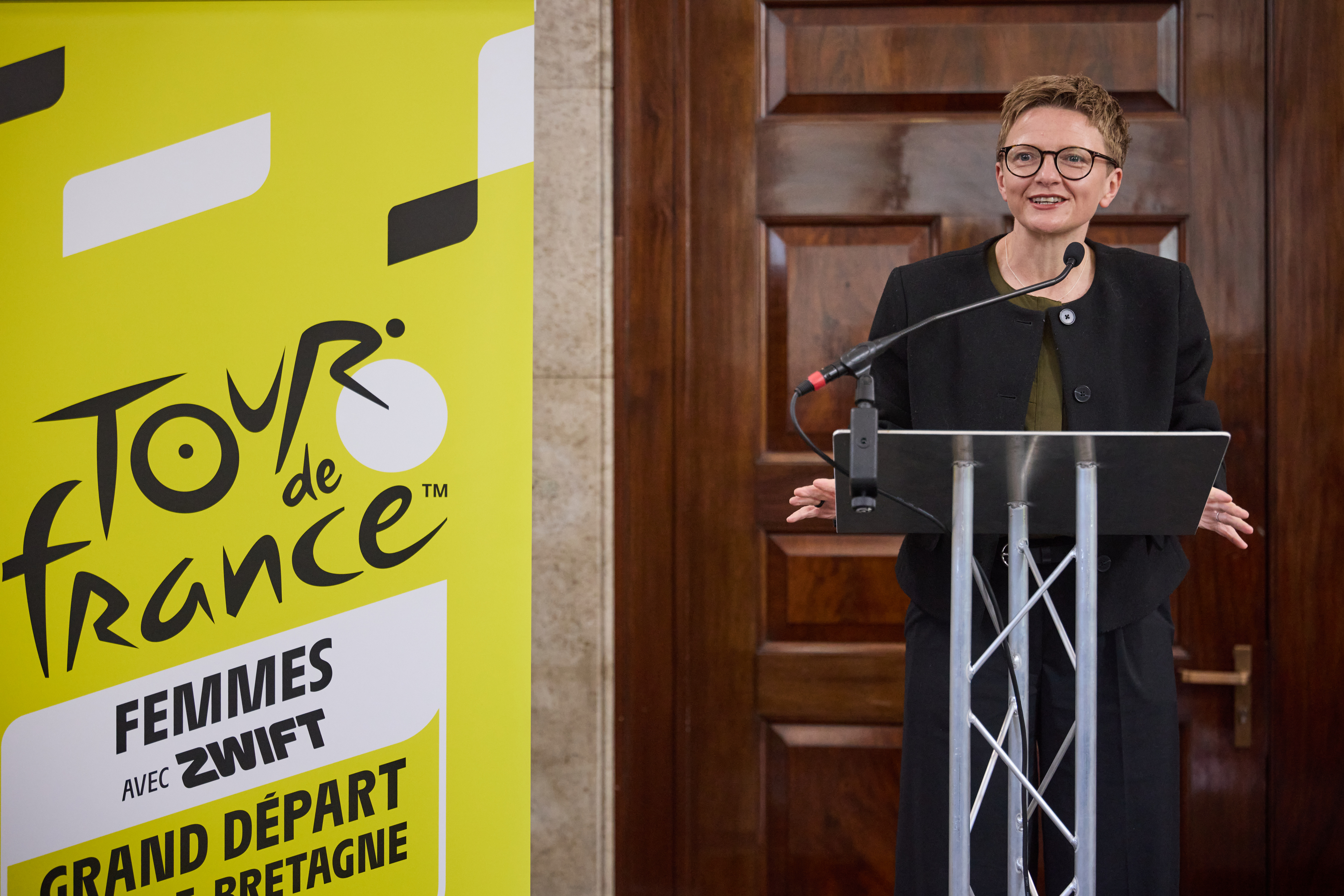Bev Craig Leader of Manchester City Council speaking from a stage with a yellow banner to one side saying Tour de France Femmes