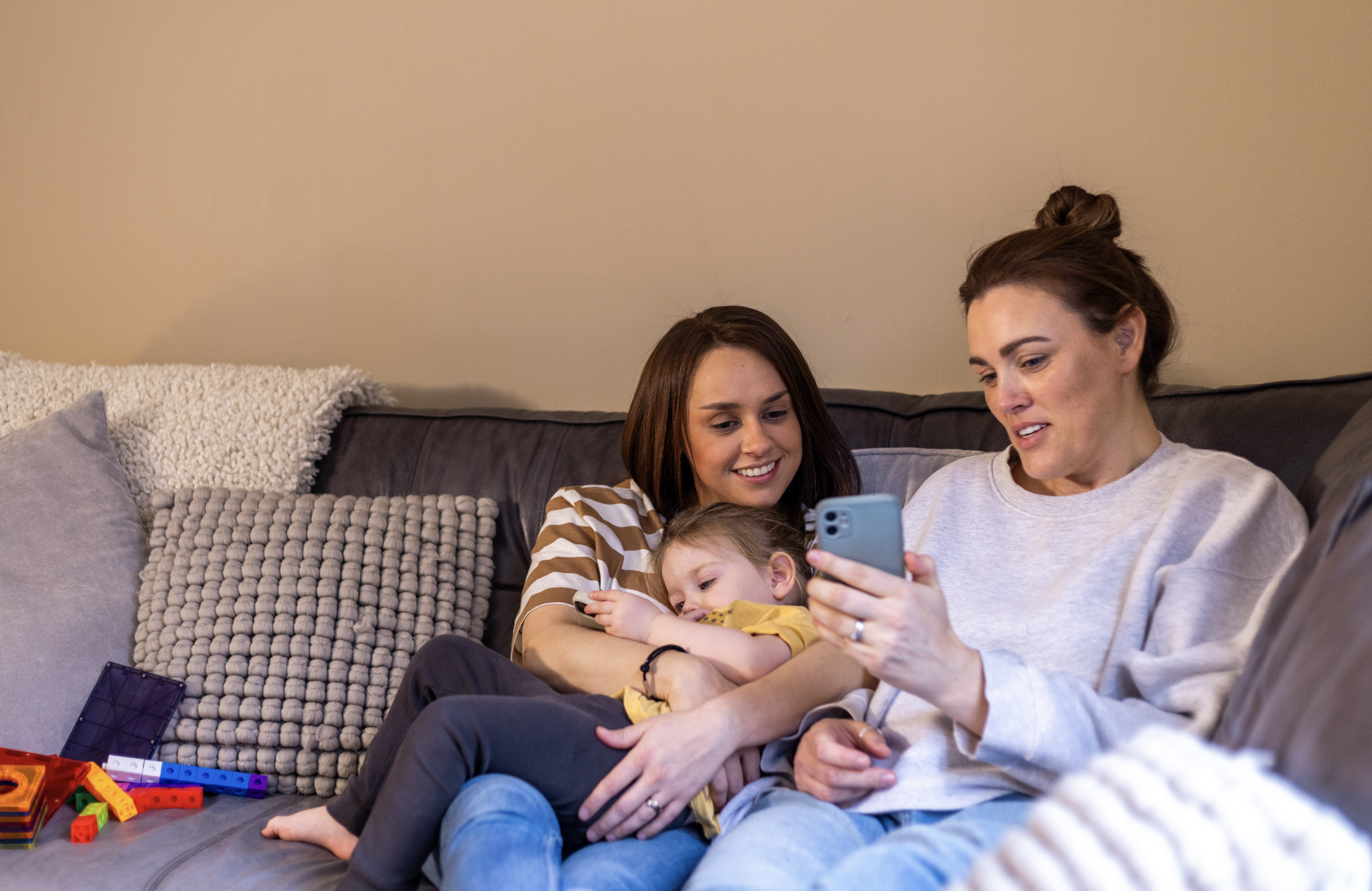 A generic picture of two women sat on a sofa with a small child, looking at a phone.