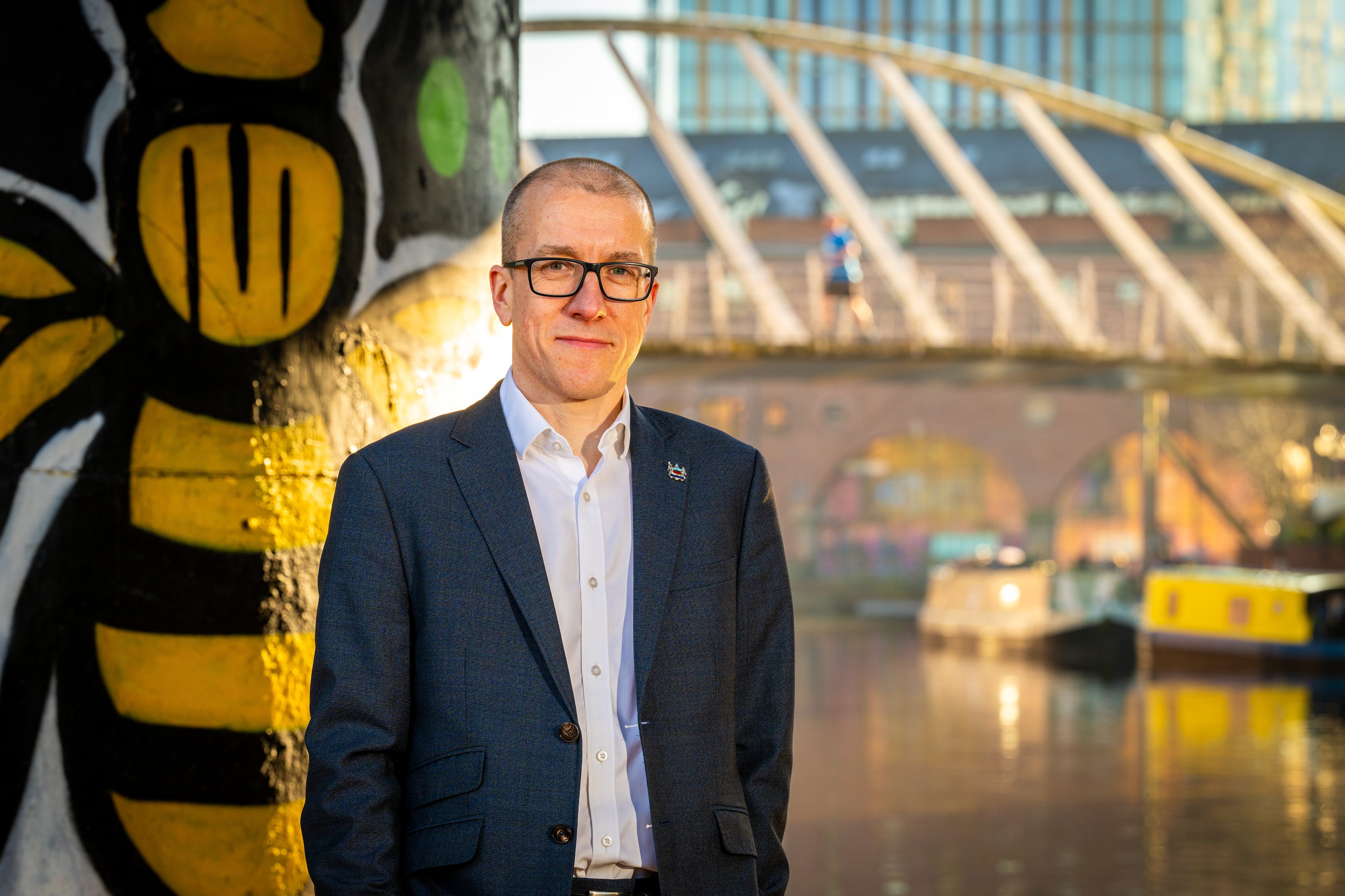 Man wearing glasses, a shirt and dark jacket, looking at camera, standing in front of a river with a barge on it and a bridge going across it.
