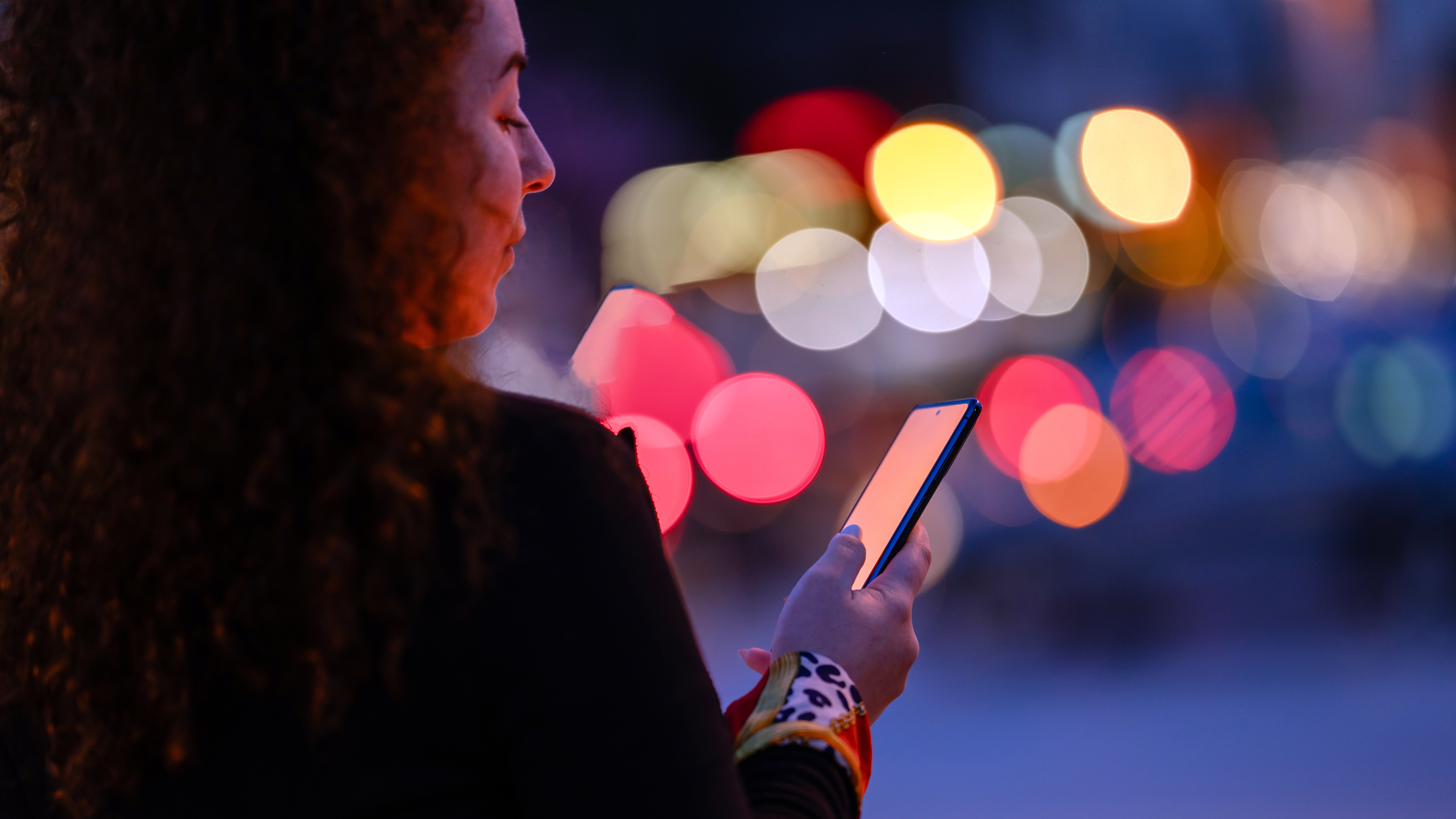Image of a woman looking down at her phone, against an illuminated night-time backdrop.