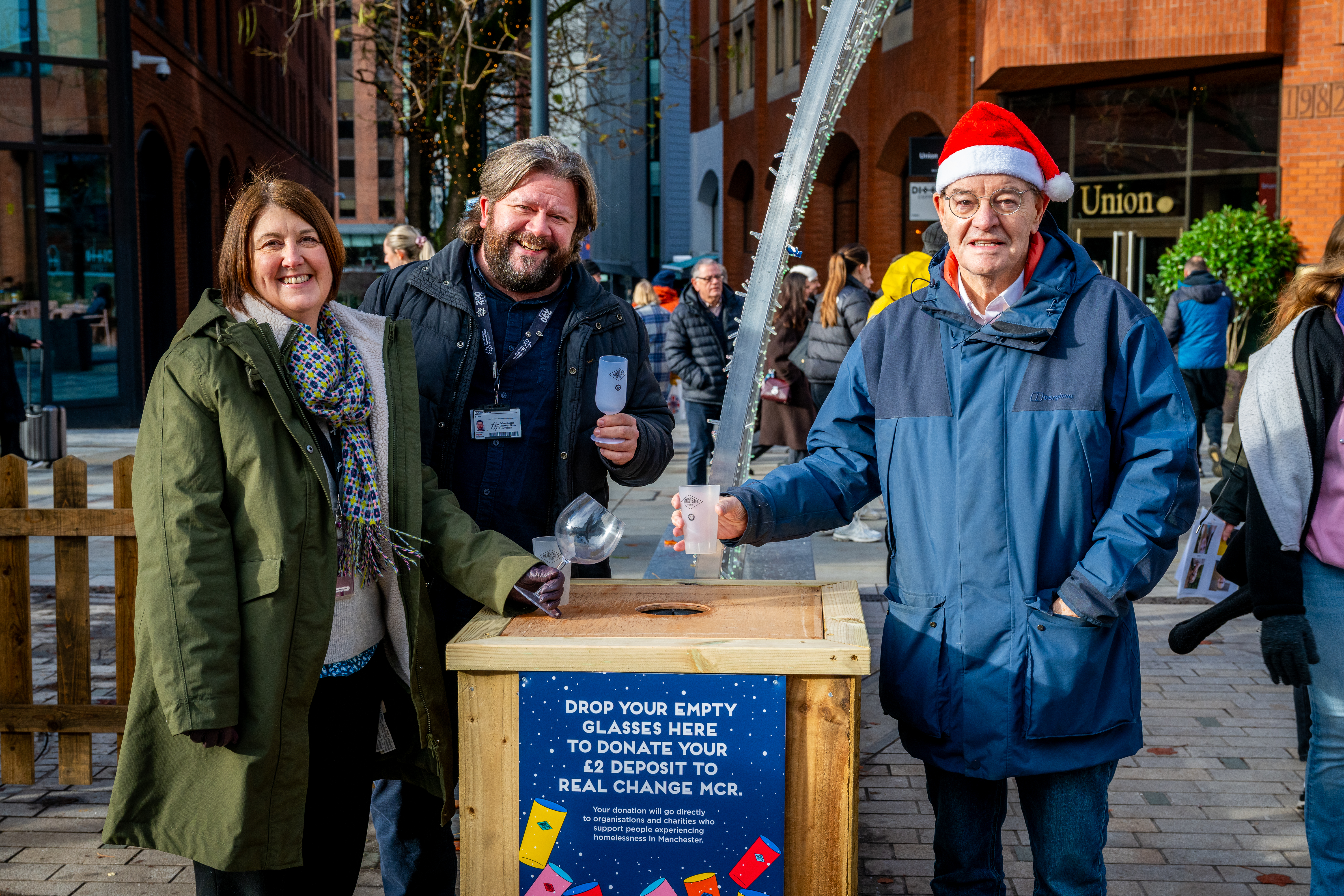 3 people stand outside around a wooden box shaped bin with the words drop your empty glasses here to donate your £2 deposit to Real Change MCR.  The people are Councillor Joanna Midgley, James Charnock and Pat Karney