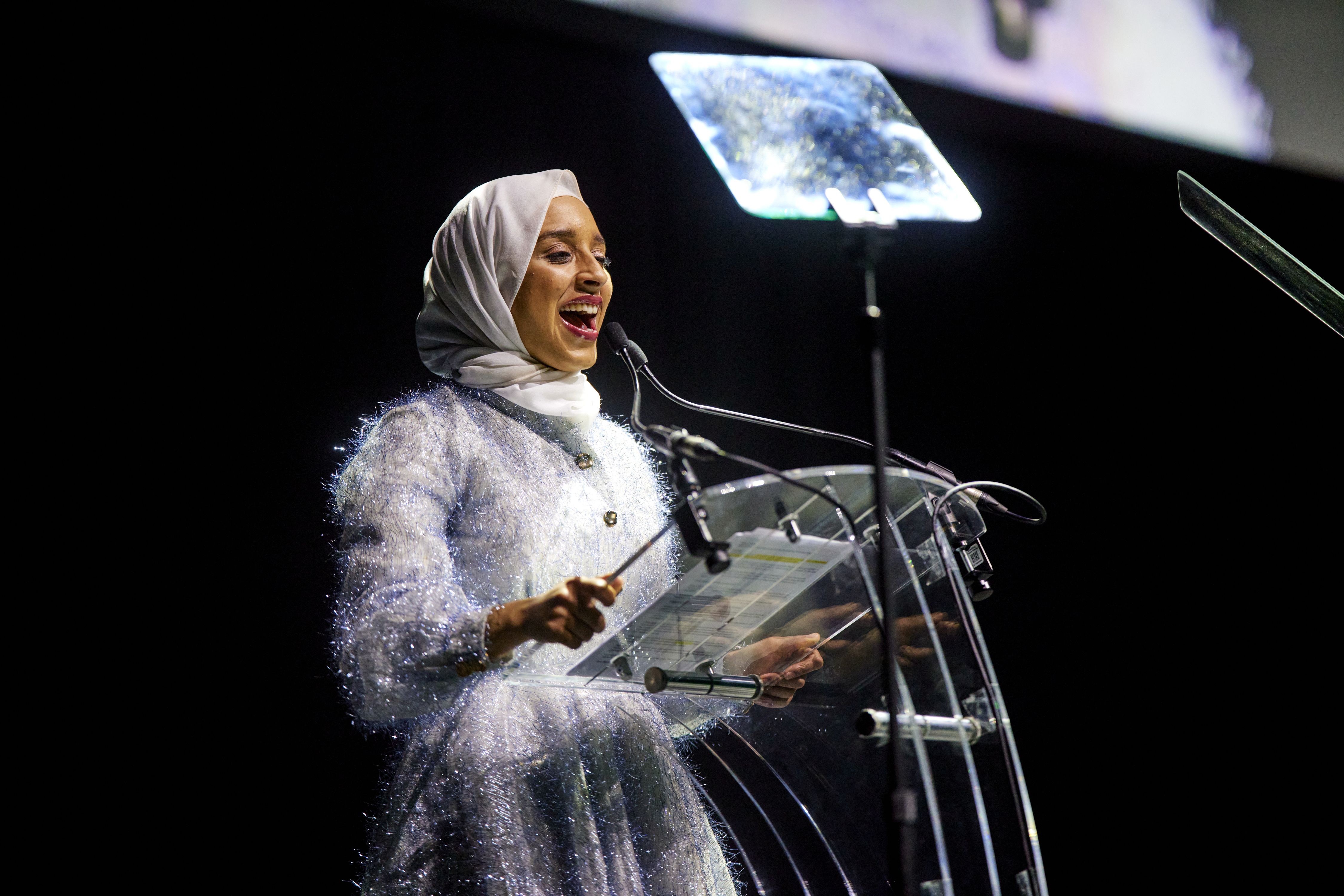 Young woman dressed in silver with a silver headscarf standing in front of a lecturn smiling as she speaks to an audience we can't see