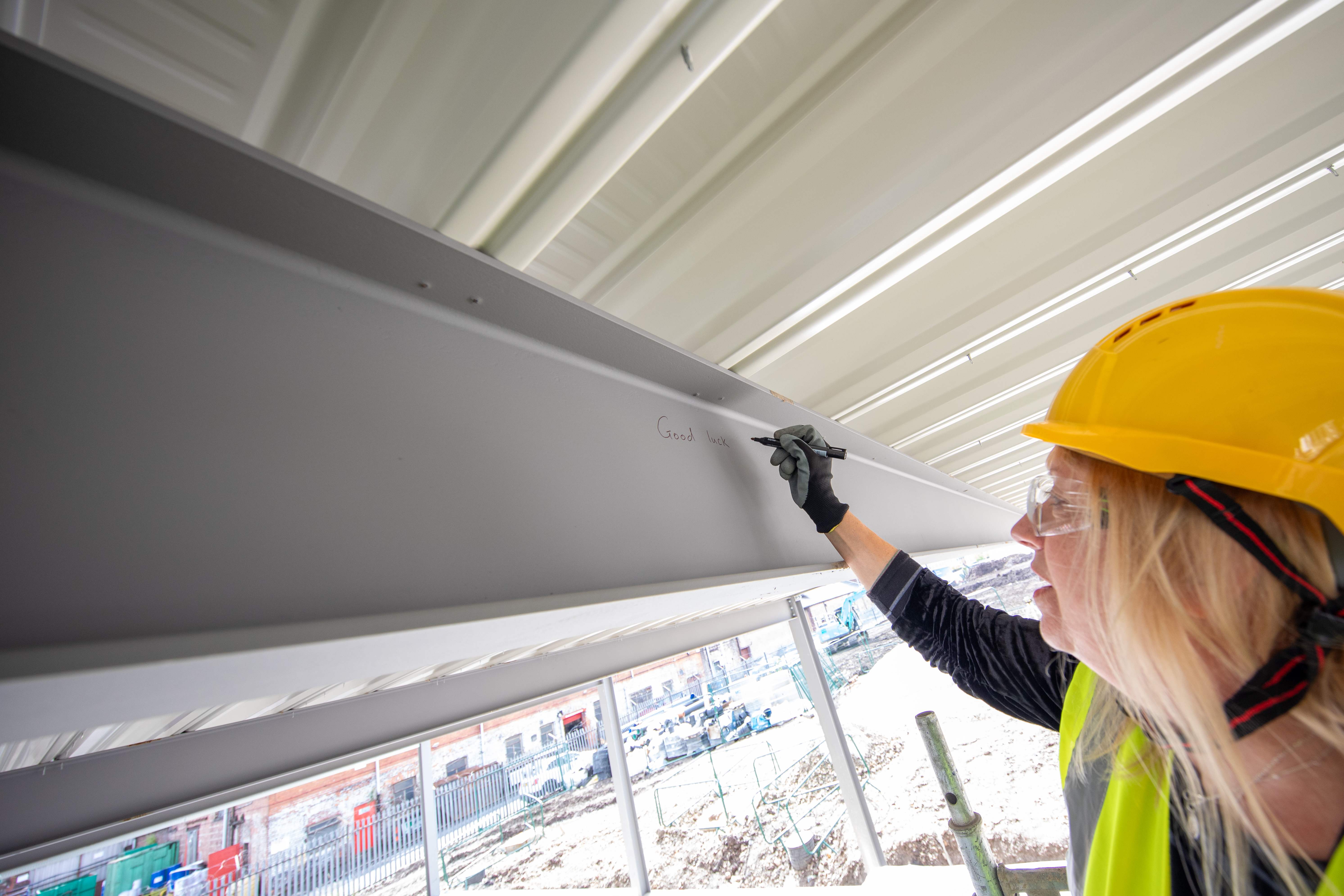 A person in a yellow hard hat is pictured writing on a steel beam with a pen A person in a yellow hard hat is pictured writing on a steel beam with a pen