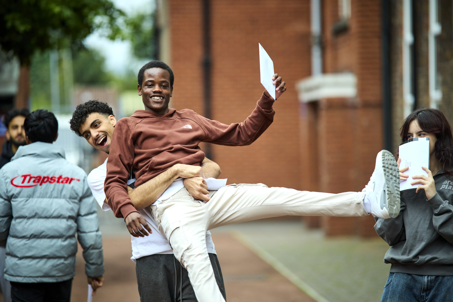 Two teenage boys both smiling as one, holding an envelope in his hand, is picked up and lifted off the ground by the other