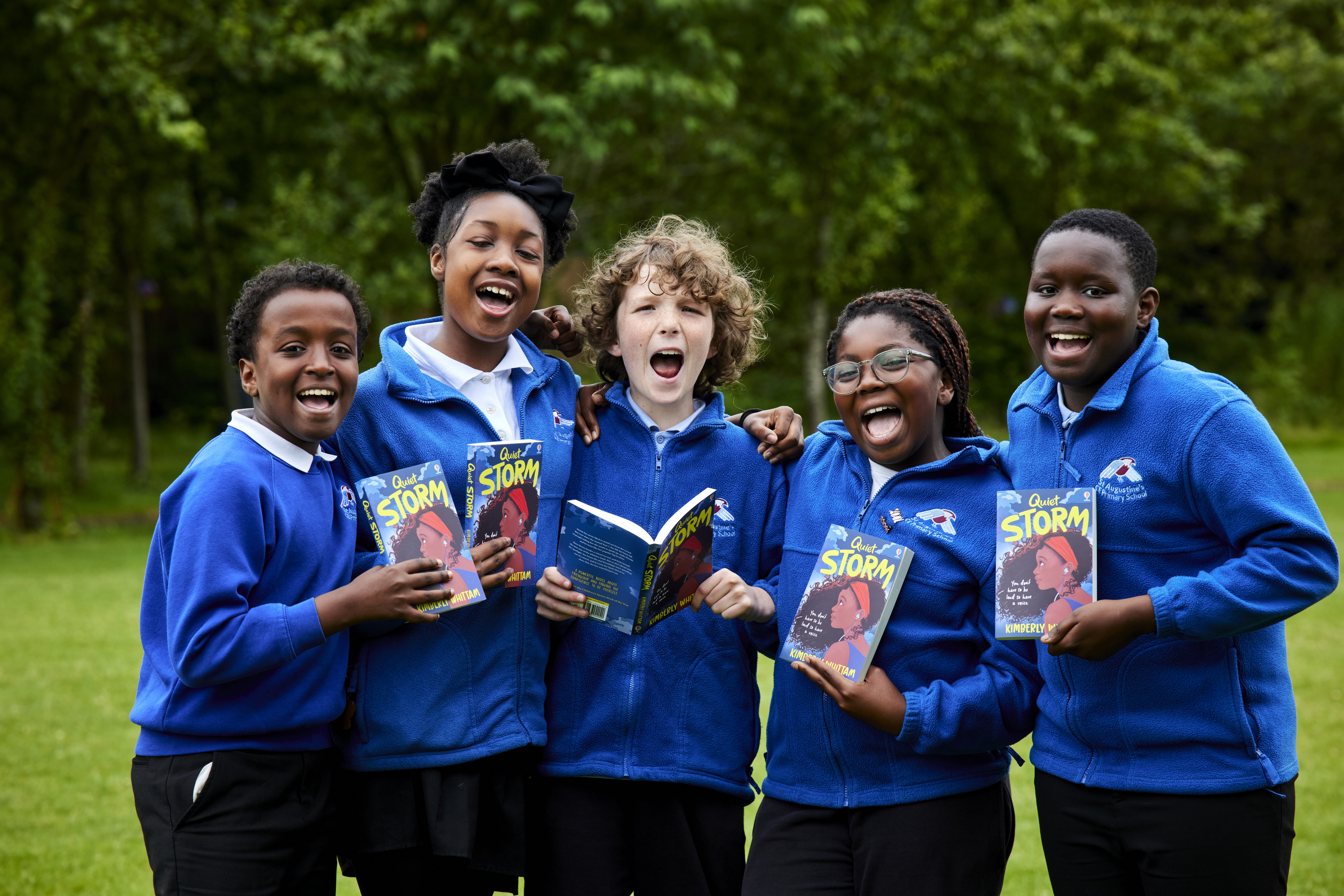 5 school pupils in blue uniforms standing close to each other, each holding a copy of the same book, smiling and shouting towards the camera