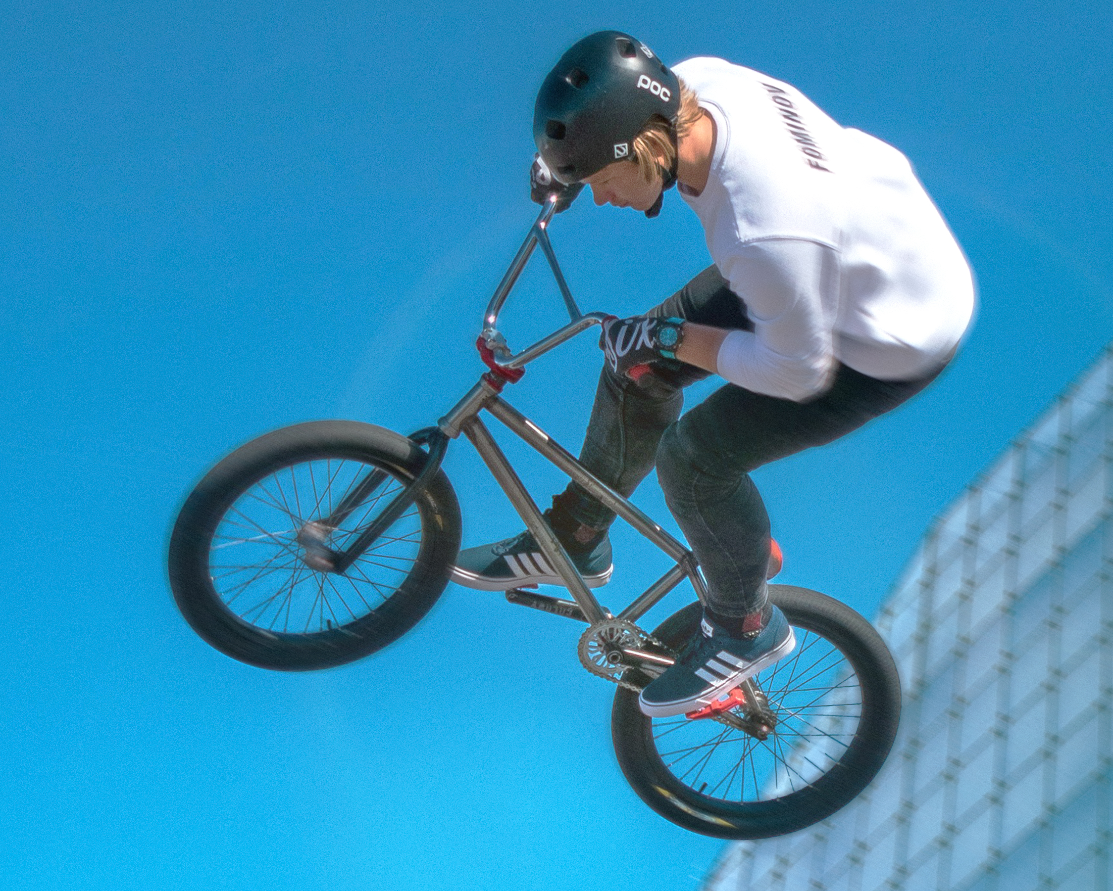 Young person on a BMX bike riding through the air against a blue sky with a glass building in the background