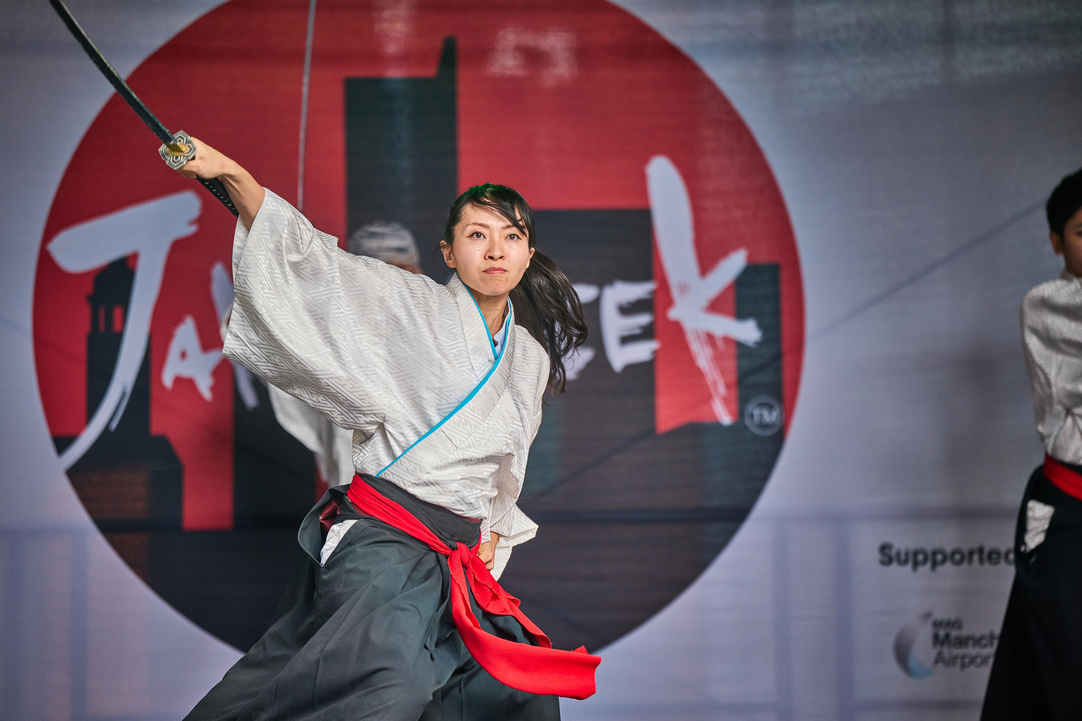 Woman dressed in traditional Samurai outfit of grey trousers and a white shirt with a red sash tied round her middle, holding a Samurai sword