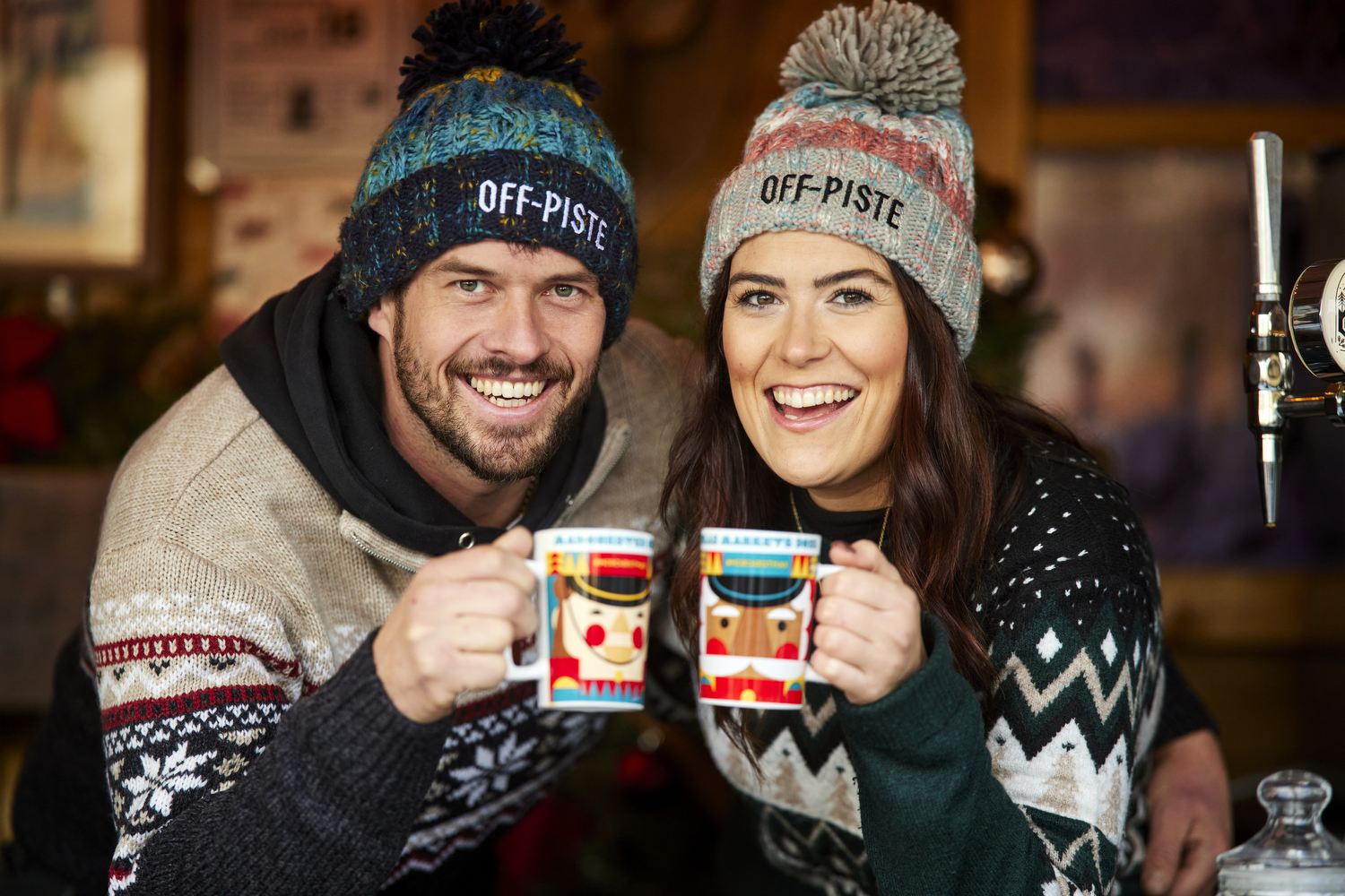 Man and woman wearing Christmas jumpers and bobble hats with the words off piste on them, hold up mugs with a nutcracker toy soldier design on them making a cheers gesture with their mugs