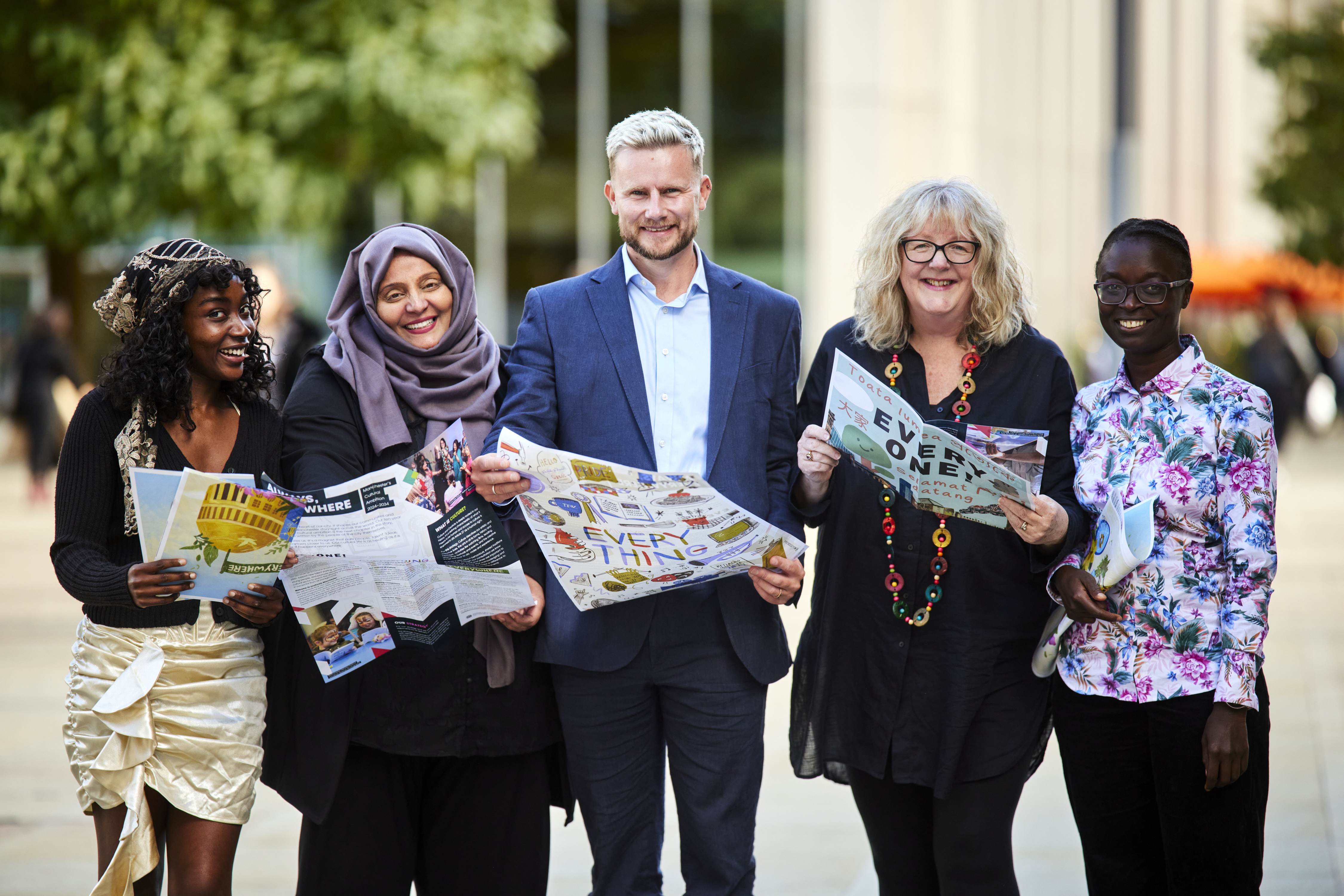 Five people standing together in a row outside in a public space smiling at the camera as they hold leaflets and posters in their hands