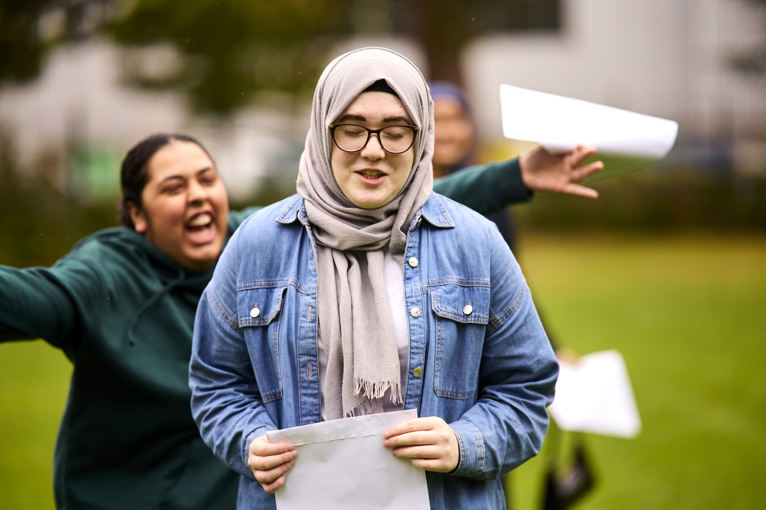 Two teenage girls, one running up behind the other smiling, arms outstretched reaching to grab the girl in front who is holding an envelope